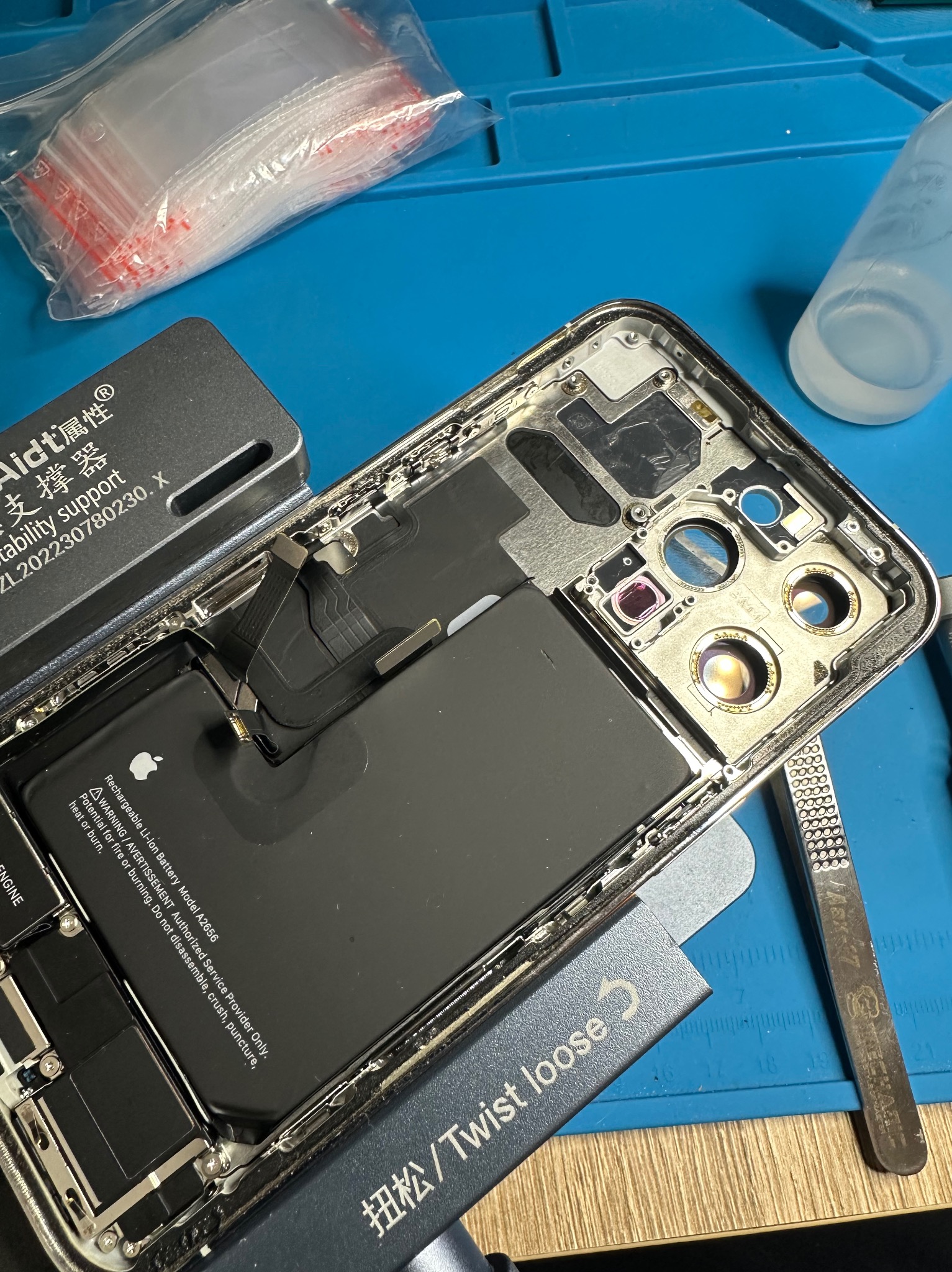 Rozłożony iPhone 13 Pro na niebieskiej macie serwisowej, widoczna bateria, narzędzia (pęseta, otwierak), elementy demontażu. Precyzyjna naprawa elektroniki.