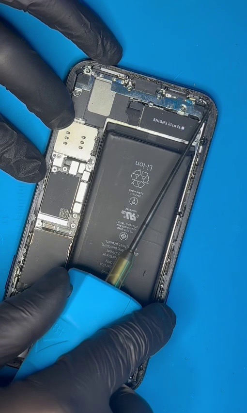 Otwarty smartfon z widoczną baterią, narzędziem i dłońmi w czarnych rękawiczkach podczas naprawy na niebieskim podłożu. Detal serwisowy.