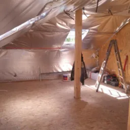 Poddasze w trakcie ocieplania, widoczne drewniane słupy, folia izolacyjna, sklejka na podłodze, drabina i narzędzia, częściowo oświetlone światłem słonecznym wpadającym przez okno dachowe.