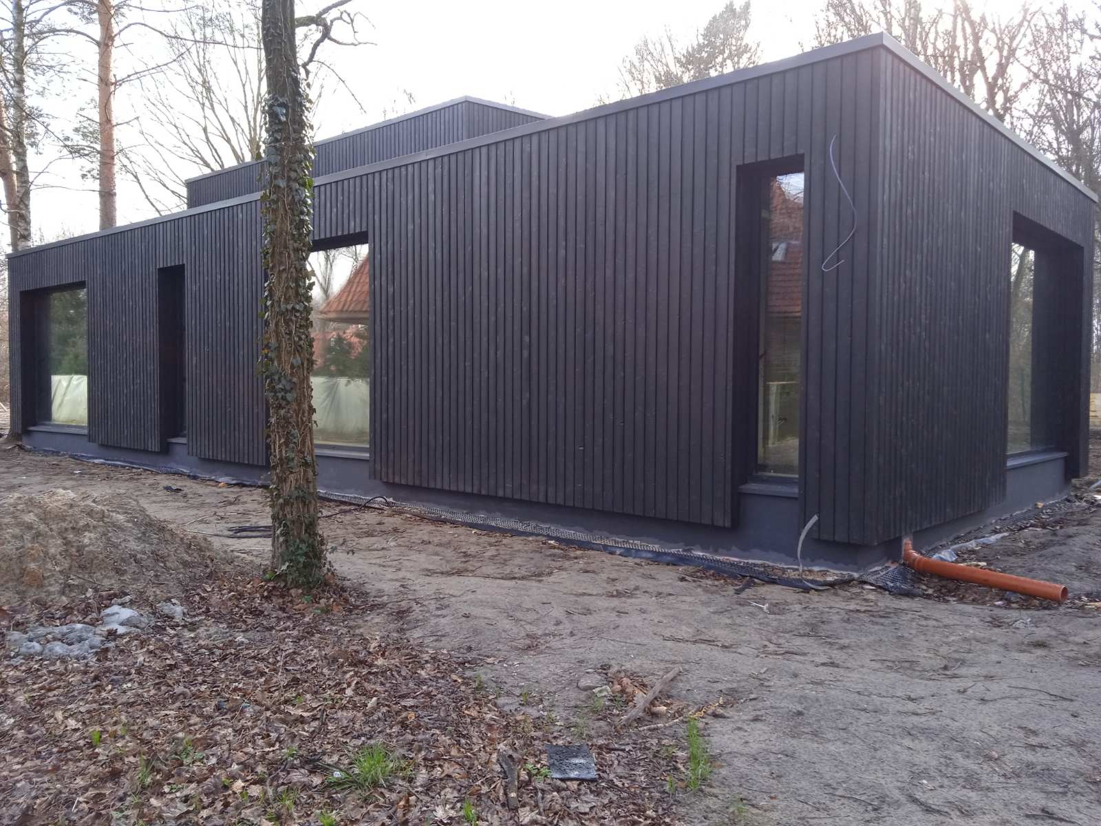 Nowoczesny dom szkieletowy z elewacją drewnianą w Otwocku, z ciemnymi, pionowo ułożonymi deskami i dużymi oknami, widoczny z zewnątrz w trakcie wykańczania terenu.