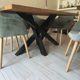 X-Weld - Stół z drewnianym blatem i metalową, krzyżową podstawą w stylu industrialnym. Krzesła tapicerowane w odcieniach szarości. Widok z niskiej perspektywy, podkreślający konstrukcję.