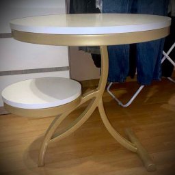 X-Weld - Złoty, metalowy stolik pomocniczy z dwoma okrągłymi blatami w kolorze kremowym, na drewnianej podłodze. Nietypowa konstrukcja nóg.