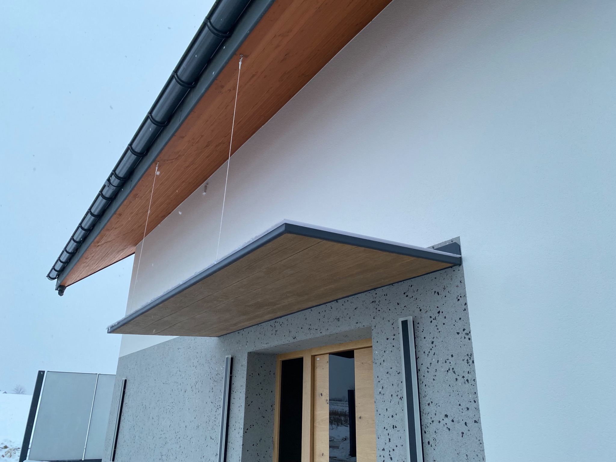 Nowoczesne zadaszenie nad wejściem do domu z drewnianym wykończeniem i szklanym panelem, widoczne w zimowej scenerii. Eleganckie detale architektoniczne.