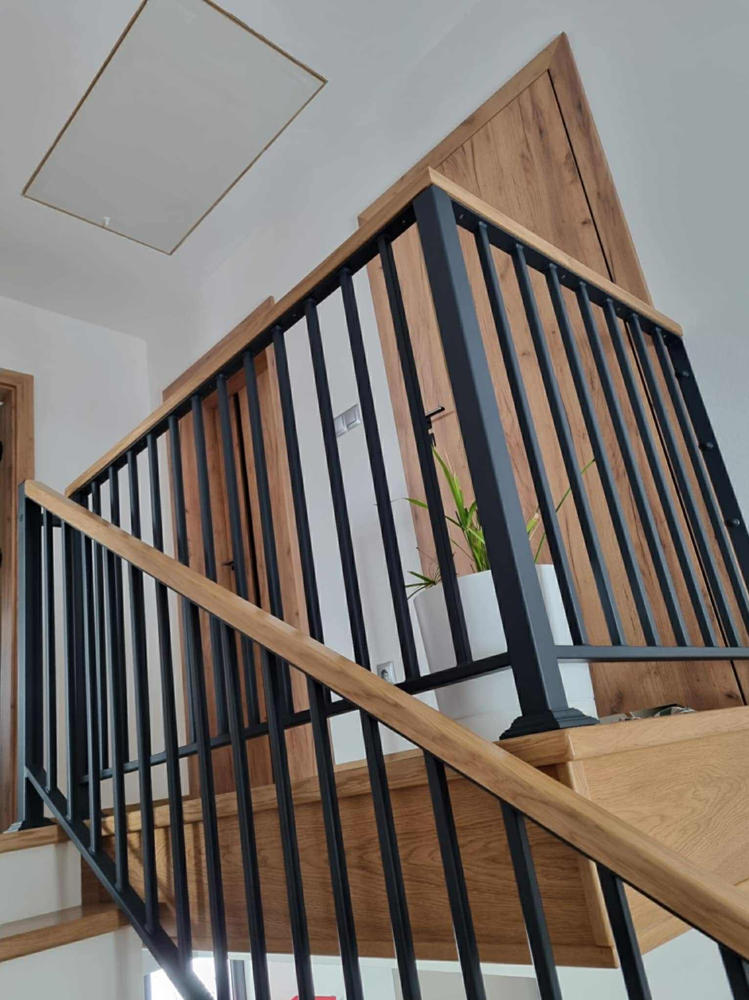Nowoczesna balustrada schodowa z czarnymi, pionowymi prętami i drewnianymi elementami, widok z dołu, roślina doniczkowa na schodach, drewniane wykończenie ściany.