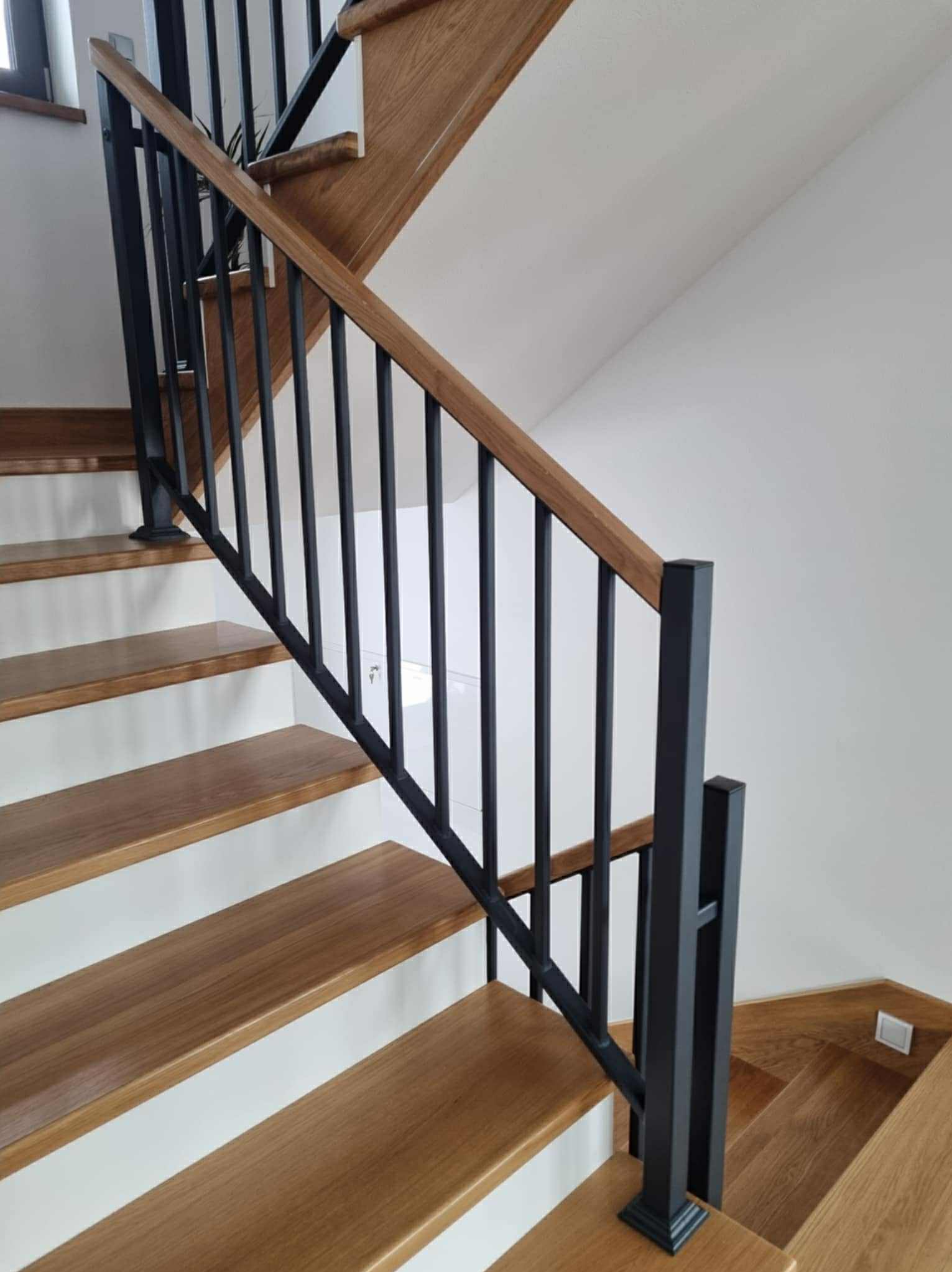 Nowoczesne schody z drewnianymi stopniami i czarną, metalową balustradą. Minimalistyczny design, jasne wnętrze. Widok z dołu, akcent na detal wykończenia.