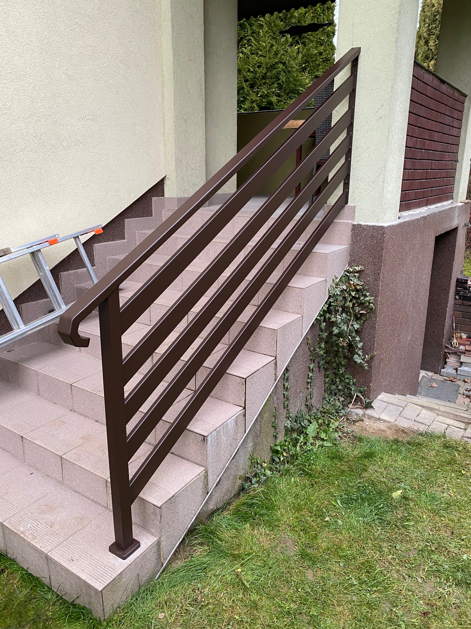 Zewnętrzne schody z brązową, metalową poręczą z poziomymi szczeblami. Widoczna zieleń trawy i pnącza. W tle ściana budynku z ceglanym elementem i drabina.