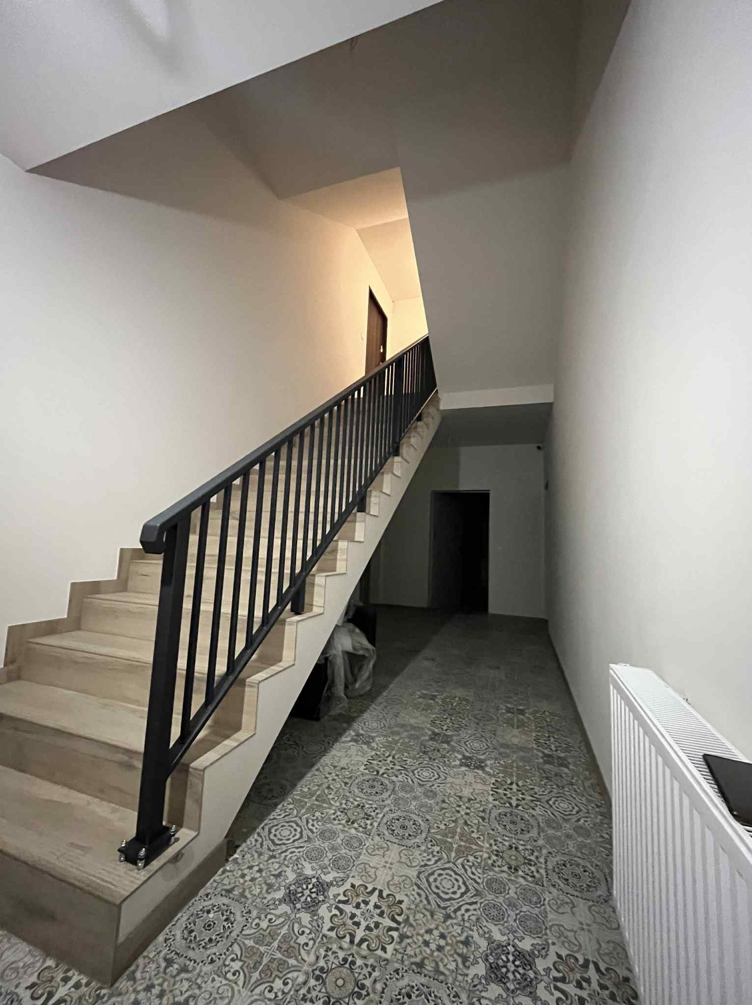 Klatka schodowa z beżowymi schodami i czarną, metalową poręczą. W tle widoczne drzwi i wzorzysta podłoga. Ujęcie z dołu, podkreślające wysokość.