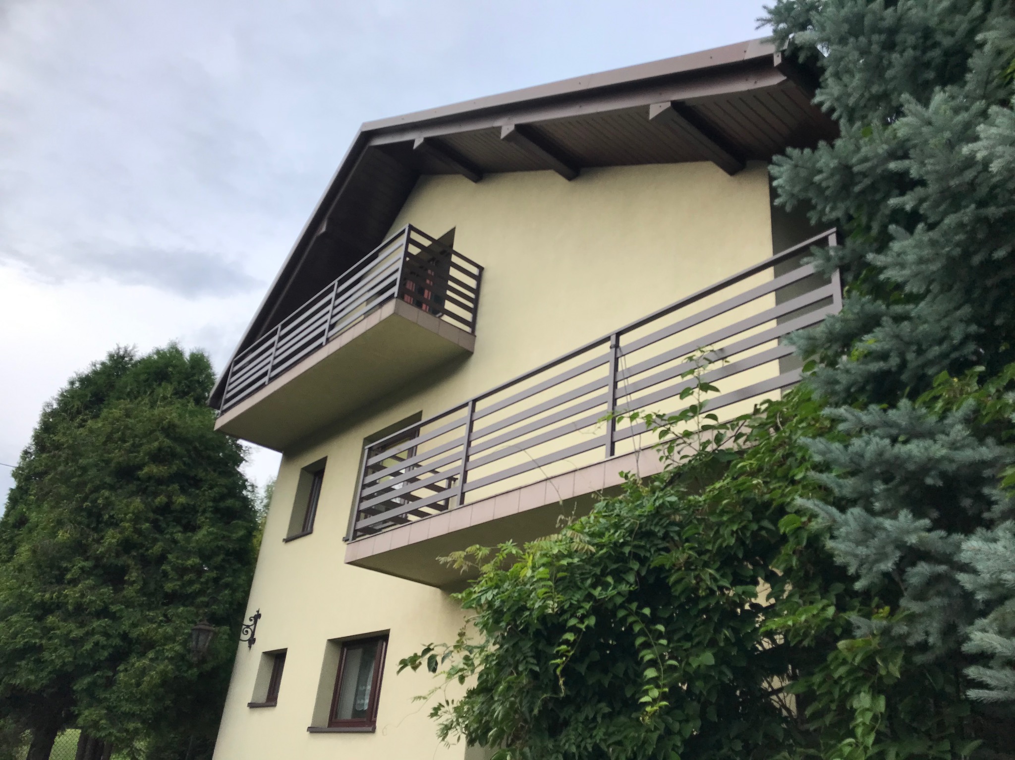 Dwupiętrowy dom z beżową elewacją i ciemnymi balustradami balkonowymi, otoczony zielenią drzew i krzewów. Ujęcie z dołu, pochmurne niebo w tle.