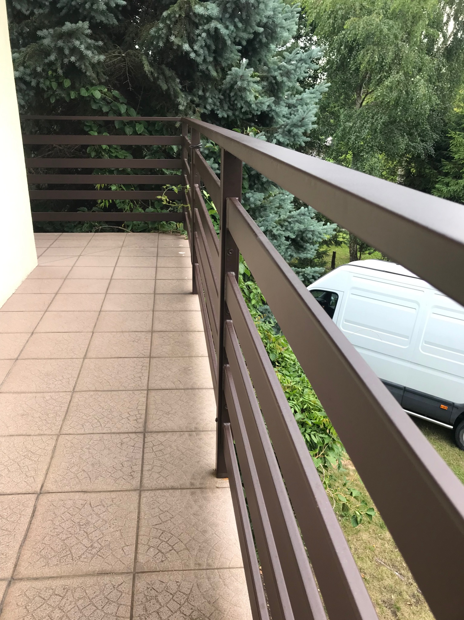 Balkon z brązową, metalową balustradą na tle zieleni i białego samochodu dostawczego. Widok z boku, ukazujący detale konstrukcji i otoczenie.