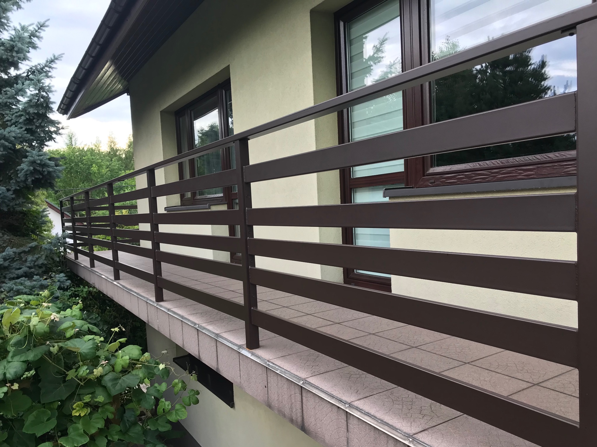 Balkon z brązową, metalową balustradą o poziomym układzie szczebli, widok z dołu, zieleń w tle. Balustrada zamontowana na balkonie wykończonym płytkami.