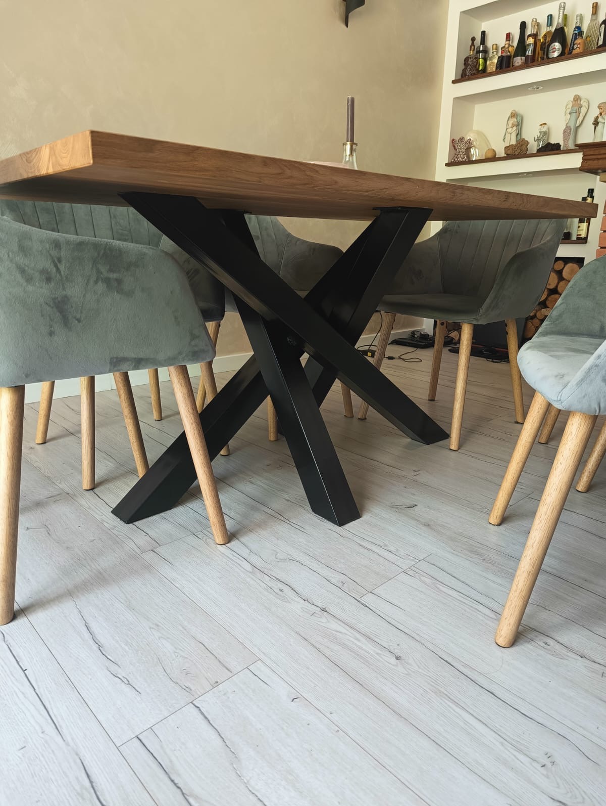 Stół z drewnianym blatem i metalową, krzyżową podstawą w stylu industrialnym. Krzesła tapicerowane w odcieniach szarości. Widok z niskiej perspektywy, podkreślający konstrukcję.