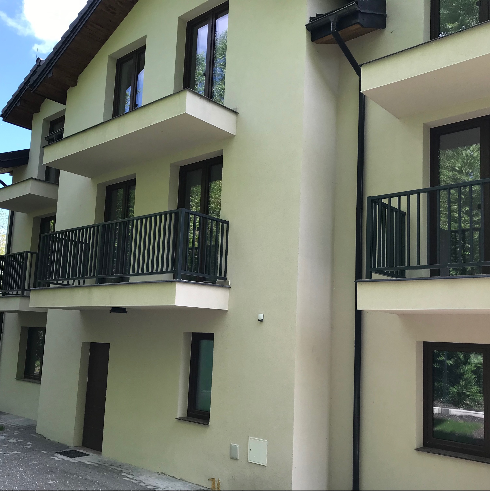 Elewacja budynku z balkonami zabezpieczonymi czarnymi, metalowymi balustradami. Widoczne okna z ciemnymi ramami i fragment dachu. Budynek w odcieniach beżu.