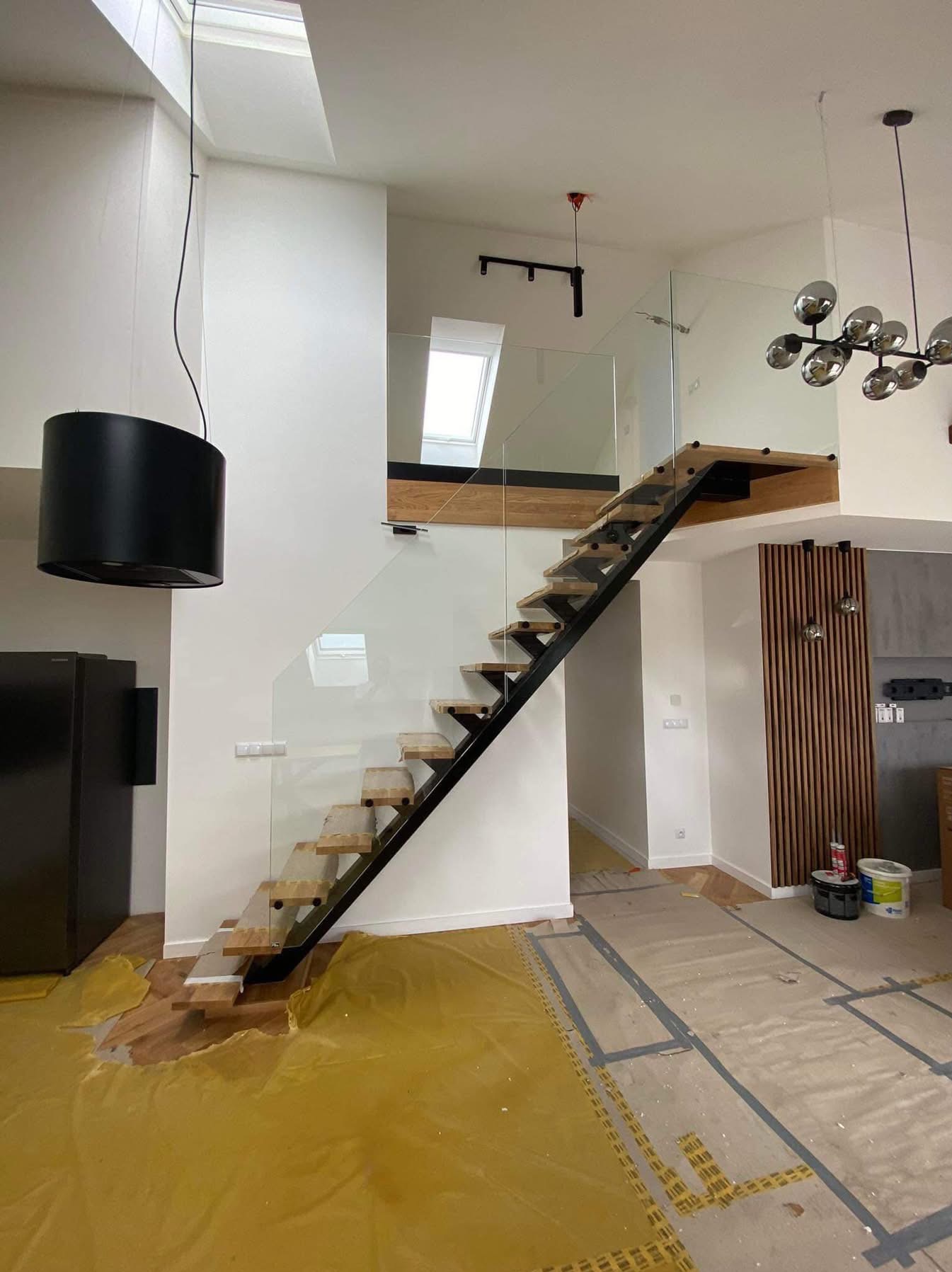Nowoczesne schody z drewnianymi stopniami i szklaną balustradą w jasnym wnętrzu. Widoczne elementy wykończenia i zabezpieczenia podłogi folią malarską.
