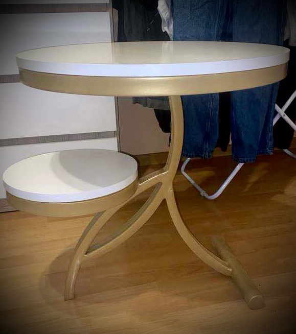 Złoty, metalowy stolik pomocniczy z dwoma okrągłymi blatami w kolorze kremowym, na drewnianej podłodze. Nietypowa konstrukcja nóg.