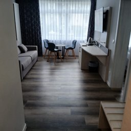 Sebastian Walko - Wnętrze pokoju hotelowego z sofą, stolikiem i krzesłami, widok na okno z zasłonami. Ciemna podłoga, jasne ściany. Widoczny kosz na śmieci i wieszak na ręczniki.