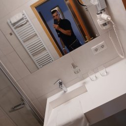 Sebastian Walko - Biała łazienka z umywalką, ręcznikiem i akcesoriami, widoczna w lustrze osoba robiąca zdjęcie. Minimalistyczny design, jasne płytki, nowoczesna armatura.