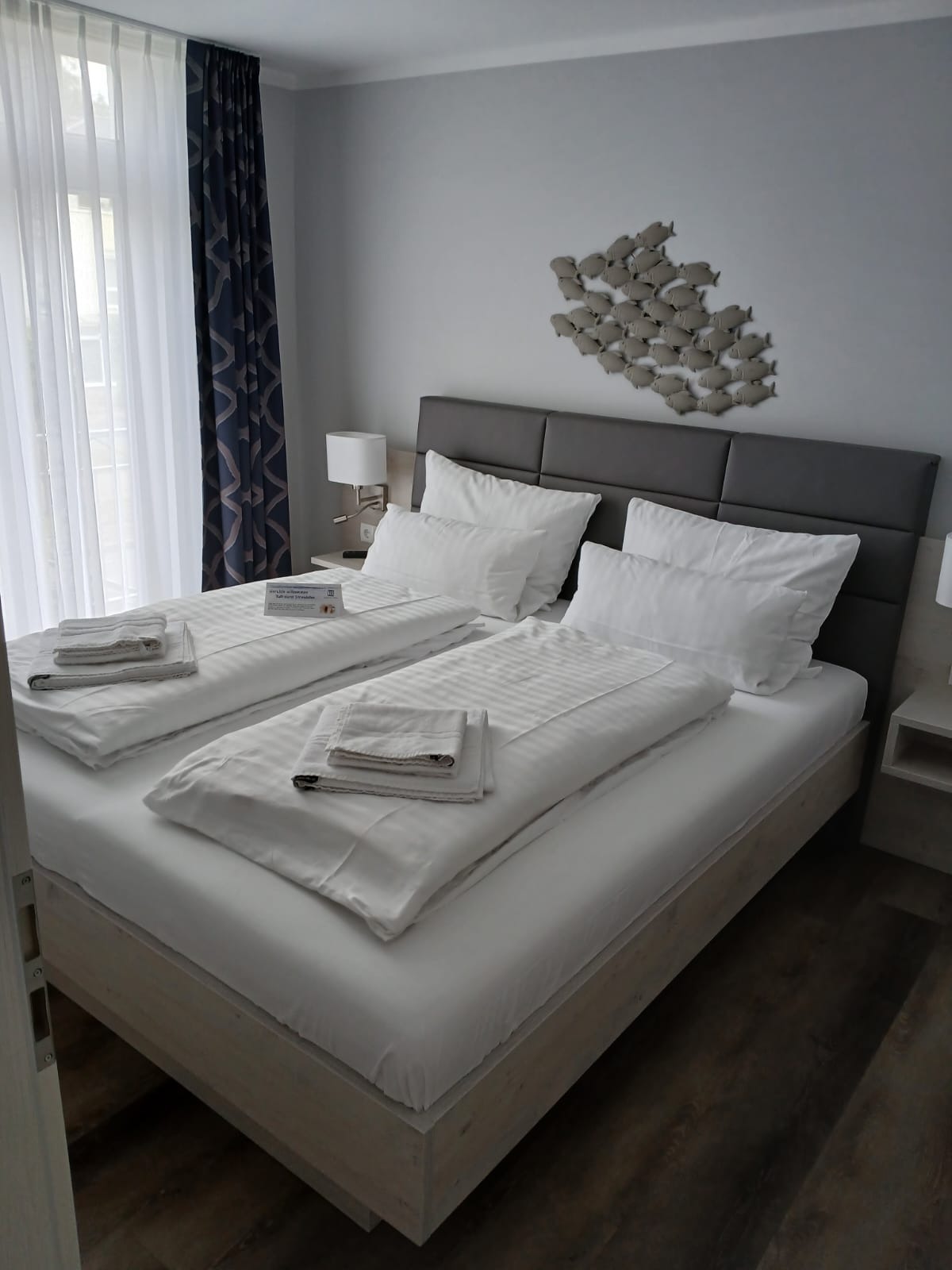 Wnętrze pokoju hotelowego z dwoma łóżkami, świeżo pościelonymi w białą pościel. Na ścianie dekoracja z ryb. Widok z perspektywy drzwi.