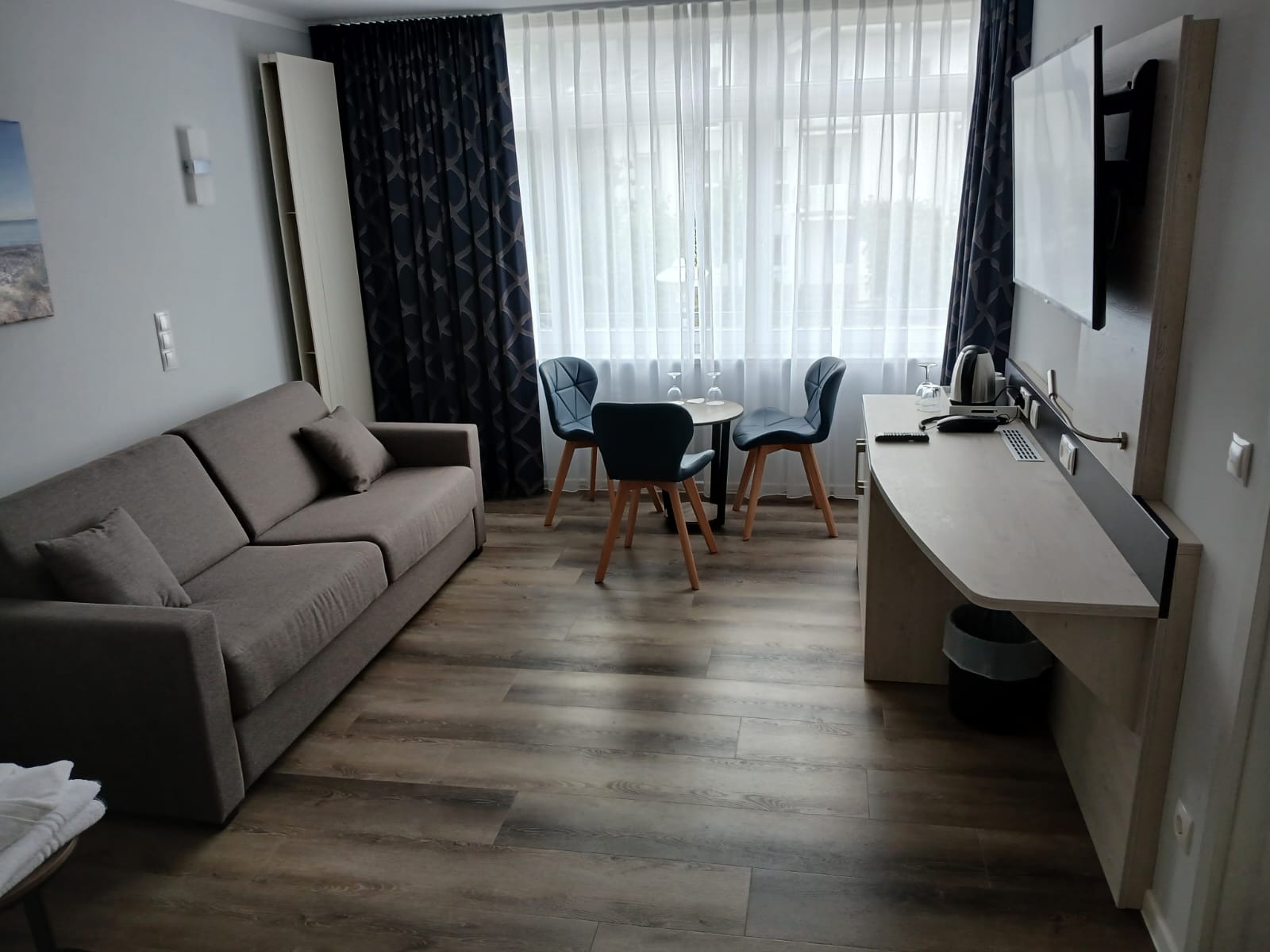 Wnętrze pokoju hotelowego z sofą, stolikiem z krzesłami, telewizorem i zasłonami. Widoczny czajnik i szklanki. Podłoga z paneli imitujących drewno.