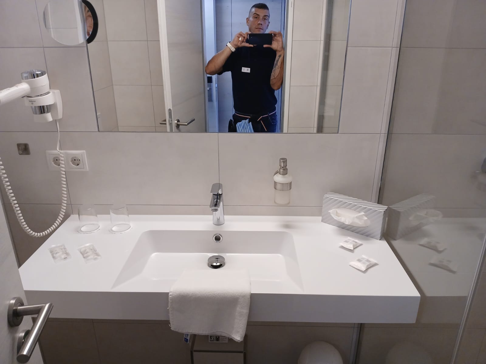 Umywalka w łazience hotelowej z akcesoriami, widok z perspektywy osoby robiącej zdjęcie w lustrze. Na blacie mydło w płynie, ręcznik i kosmetyki.