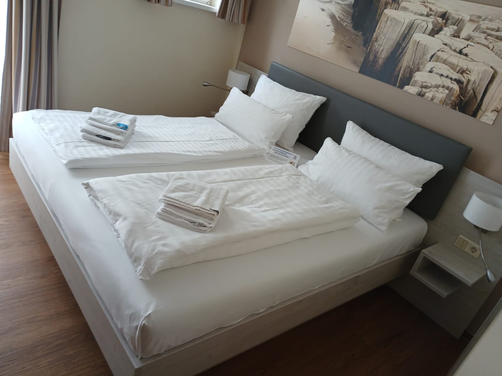 Czyste, białe pościele i ręczniki na dwóch łóżkach w pokoju hotelowym. Widoczne drewniane podłogi i obraz na ścianie. Przygotowane do przyjęcia gości.