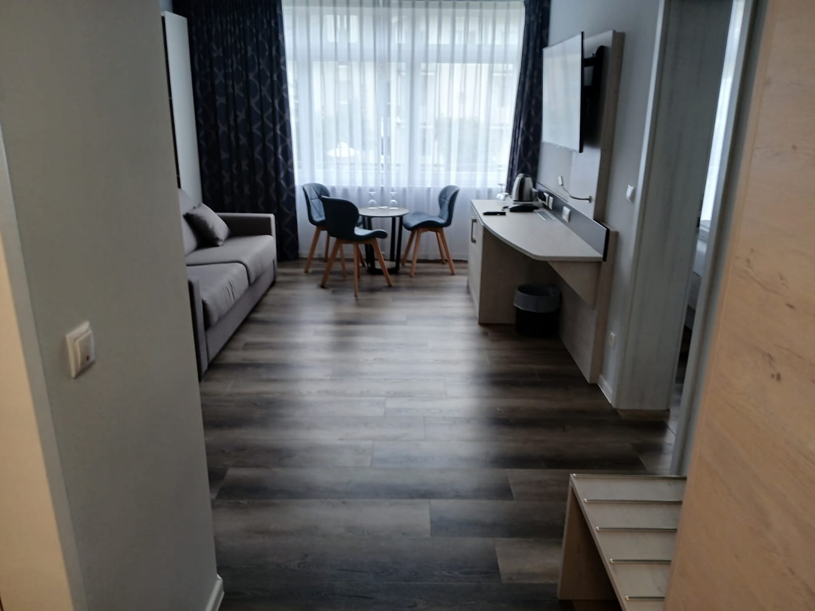 Wnętrze pokoju hotelowego z sofą, stolikiem i krzesłami, widok na okno z zasłonami. Ciemna podłoga, jasne ściany. Widoczny kosz na śmieci i wieszak na ręczniki.