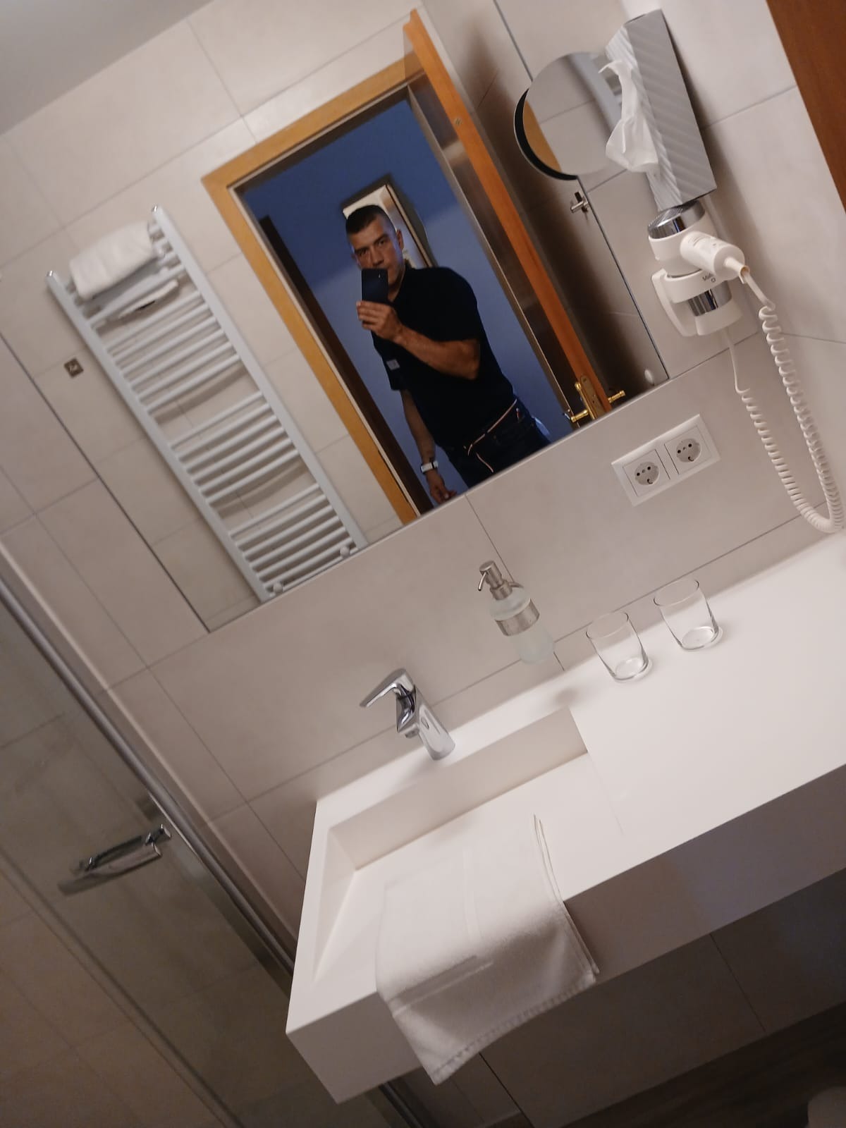 Biała łazienka z umywalką, ręcznikiem i akcesoriami, widoczna w lustrze osoba robiąca zdjęcie. Minimalistyczny design, jasne płytki, nowoczesna armatura.