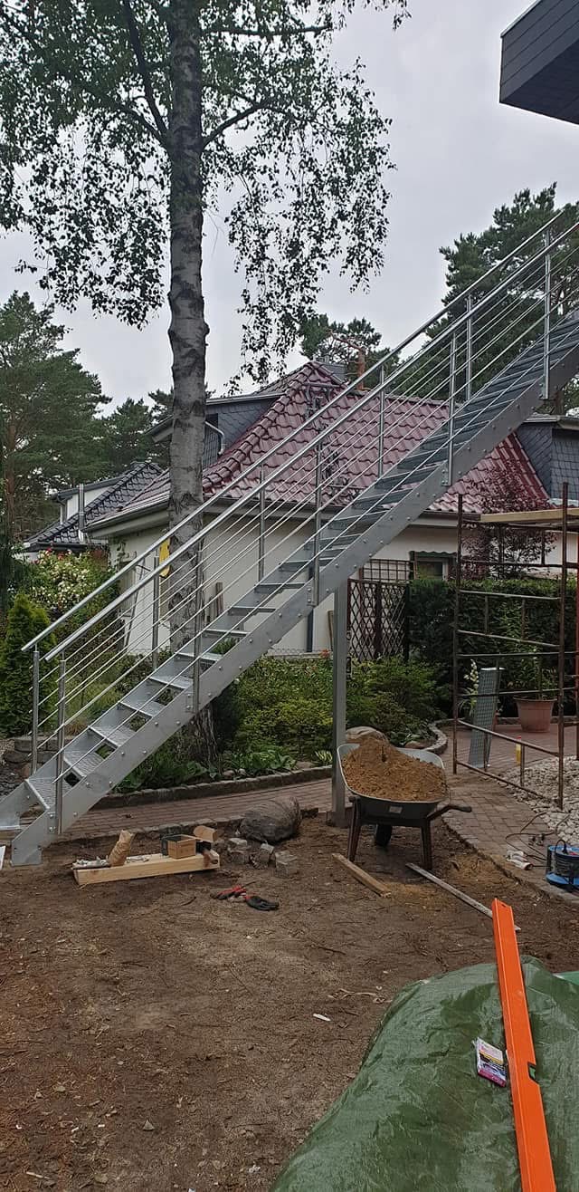 Zewnętrzne schody metalowe z poręczą, prowadzące do domu z czerwonym dachem. W tle drzewo i elementy ogrodu. Widoczne narzędzia i taczka z piaskiem.