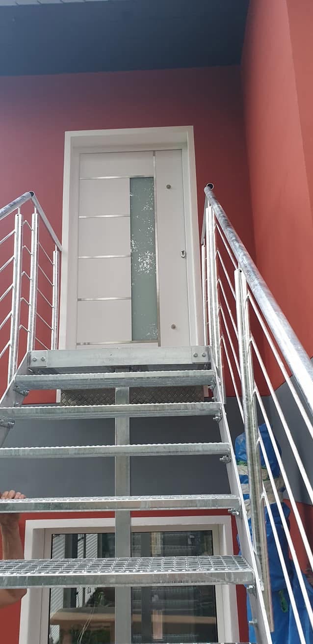 Metalowe schody zewnętrzne z poręczami prowadzące do białych drzwi z szybą, na tle czerwonej ściany budynku. Widoczne elementy montażowe.