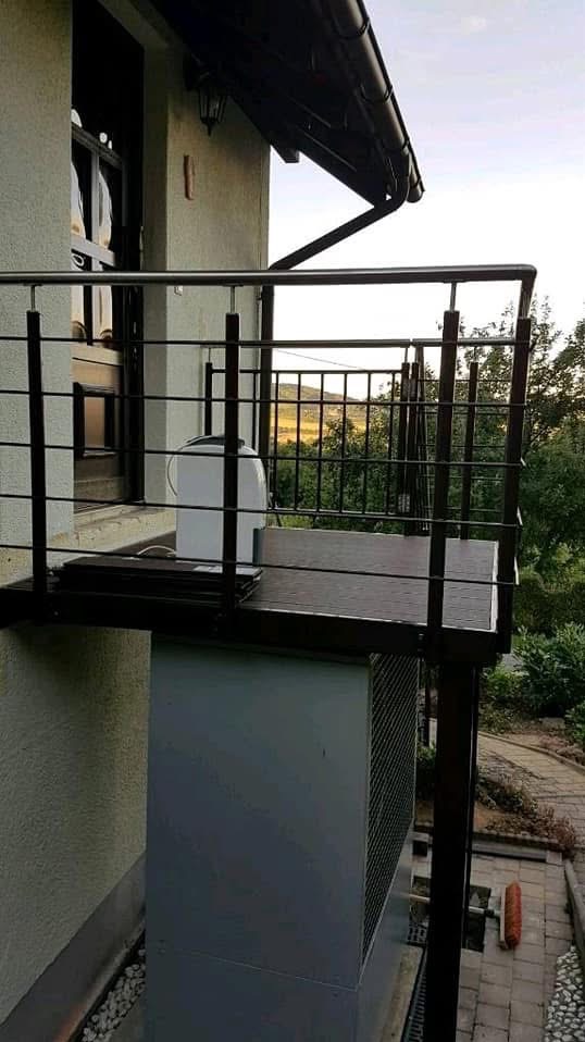 Balkon z czarną, metalową balustradą z poziomymi prętami i nierdzewną poręczą. Widok na zielony krajobraz. Na balkonie stoi biały przedmiot.