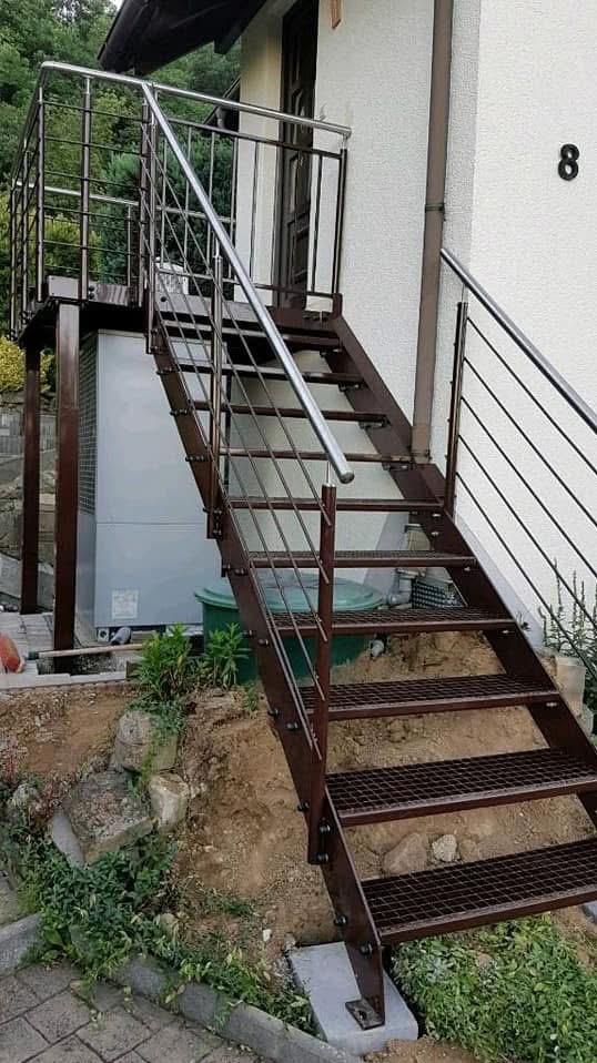 Zewnętrzne schody metalowe z brązową konstrukcją i srebrnymi poręczami prowadzące do drzwi budynku z numerem 8, otoczone zielenią i elementami krajobrazu.