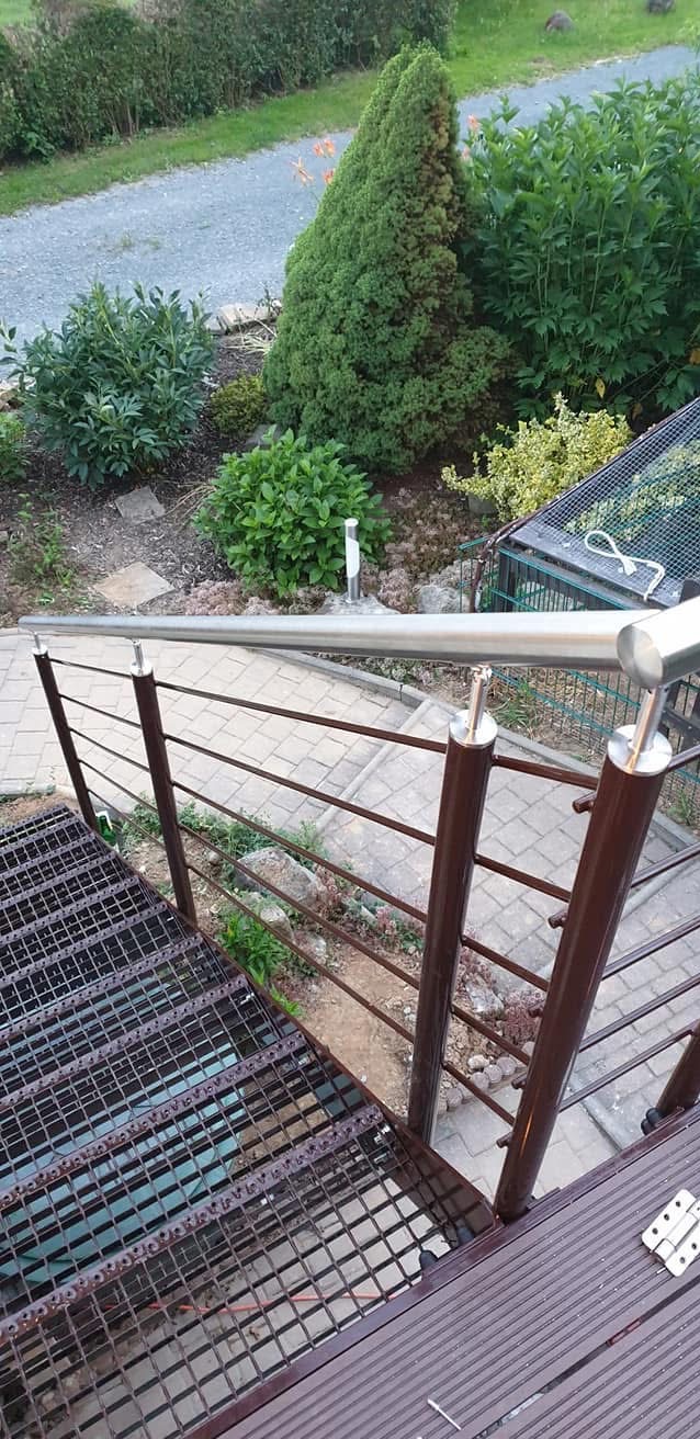 Zewnętrzne schody metalowe z brązową kratą i balustradą ze stali nierdzewnej, prowadzące do ogrodu z zielenią i ścieżką z kostki brukowej.