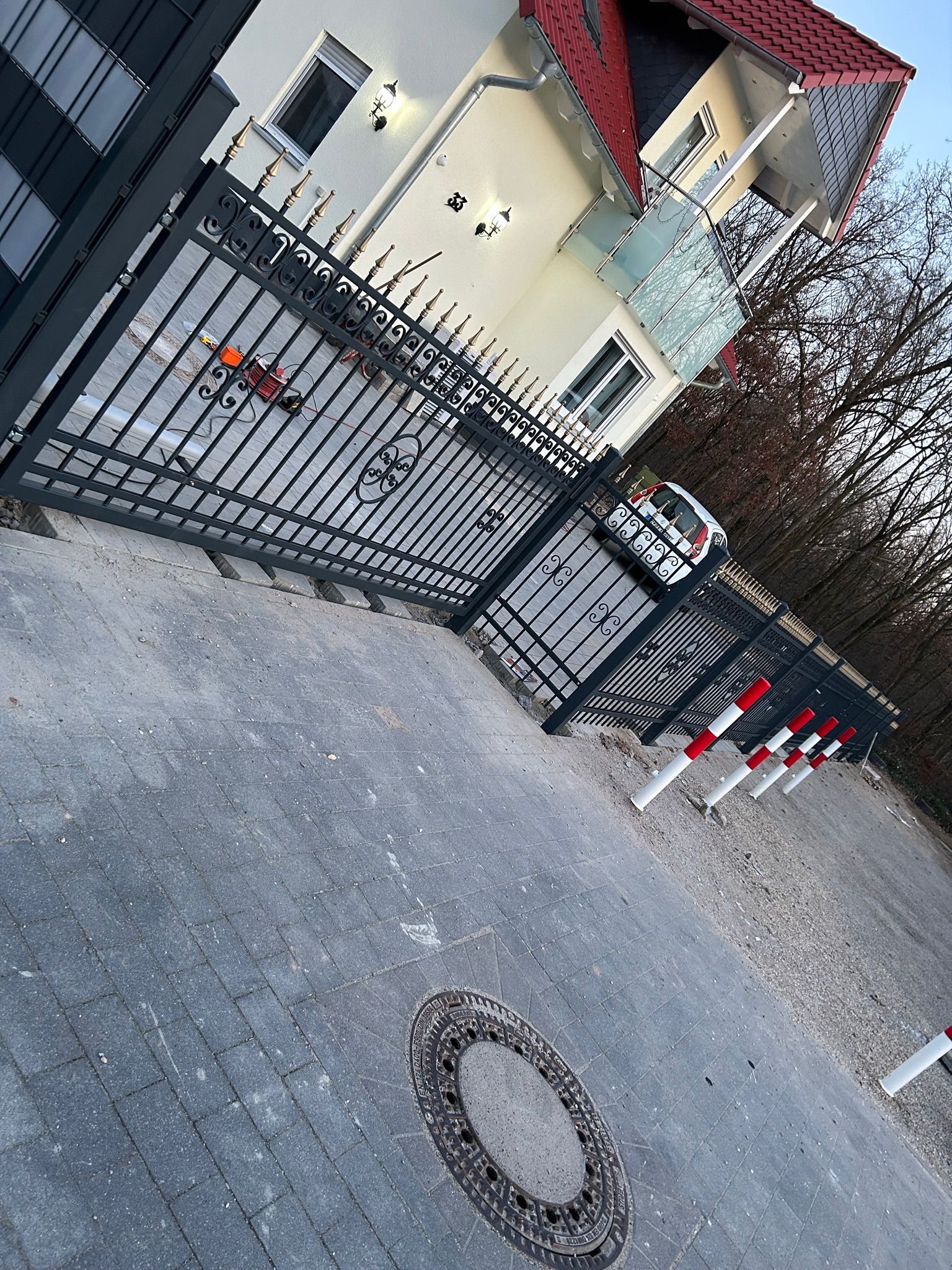 Ciemnoszare ogrodzenie metalowe z ozdobnymi elementami i złotymi grotami, otaczające posesję z domem o czerwonym dachu. Widoczne słupki z oznaczeniami.