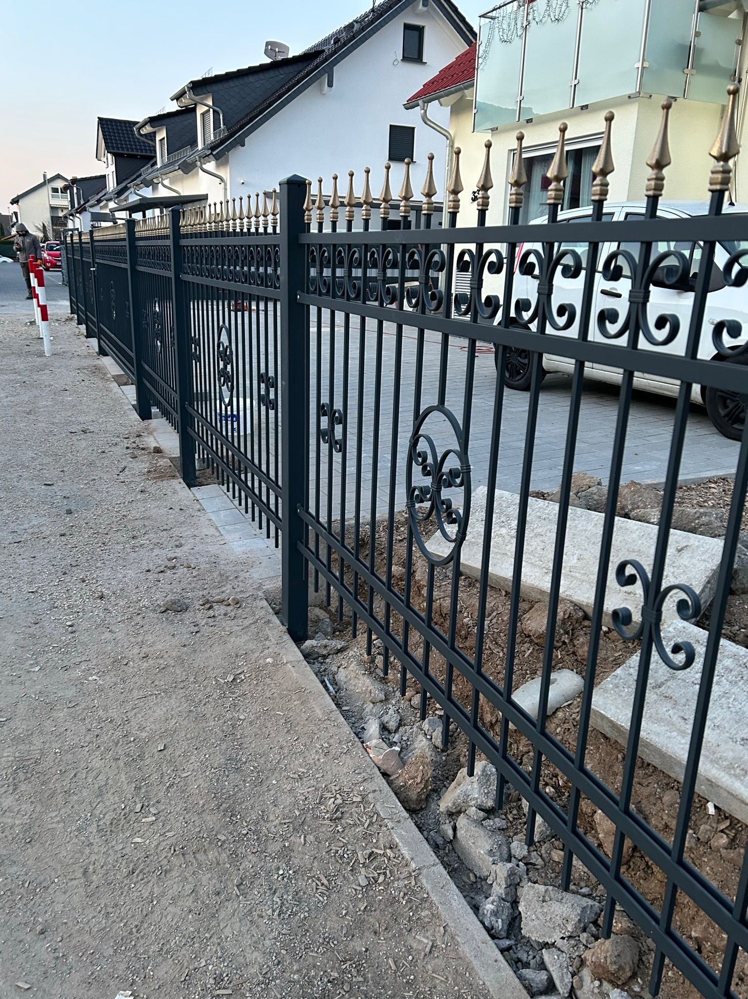 Eleganckie, czarne ogrodzenie metalowe z ozdobnymi elementami i złotymi zakończeniami, oddzielające posesję od chodnika. Widoczne domy w tle.