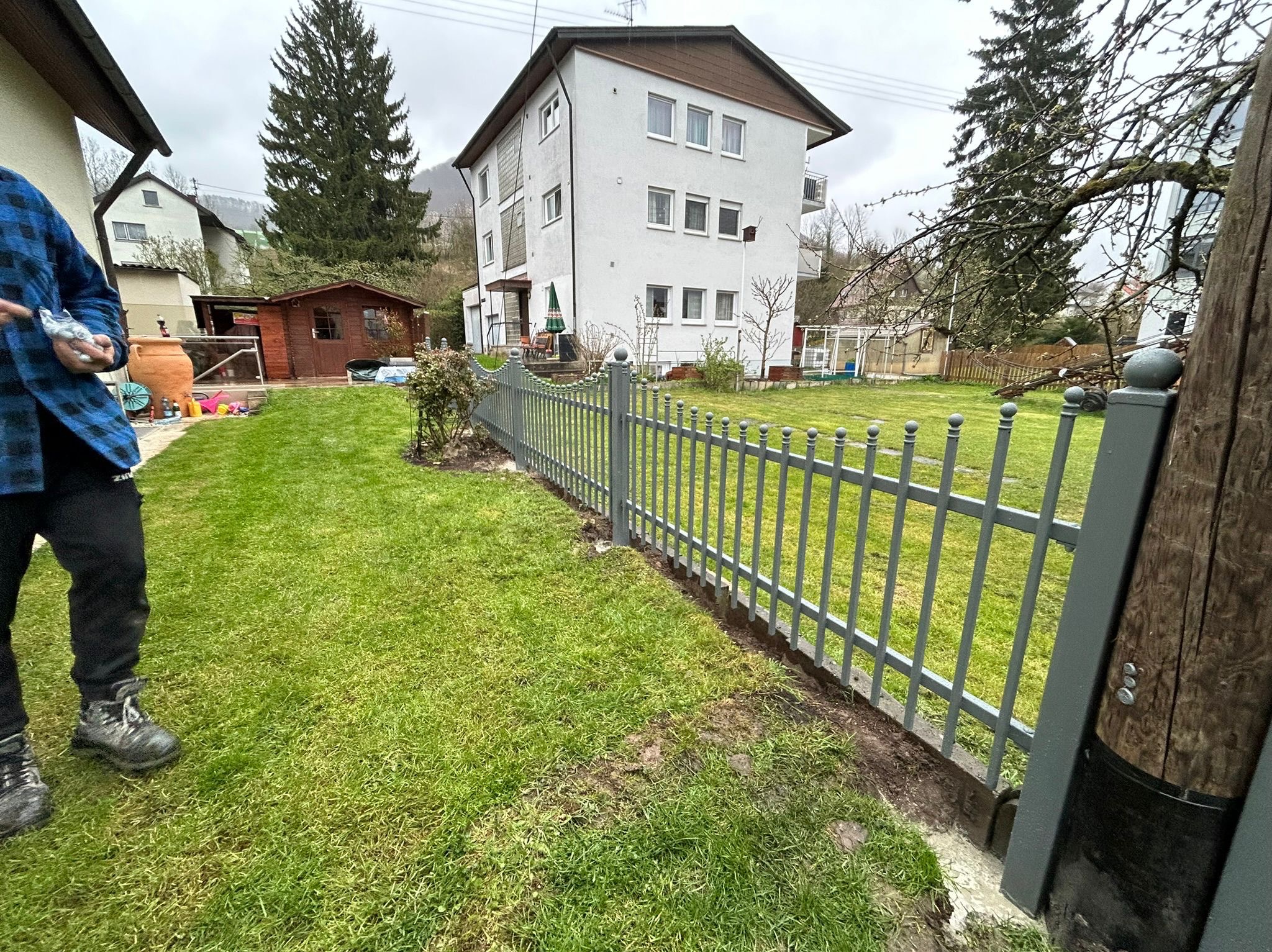 Szare ogrodzenie metalowe na trawniku przed domem. Widoczny fragment montażu, osoba w roboczych butach. Tło: budynek mieszkalny, drzewa, pochmurne niebo.