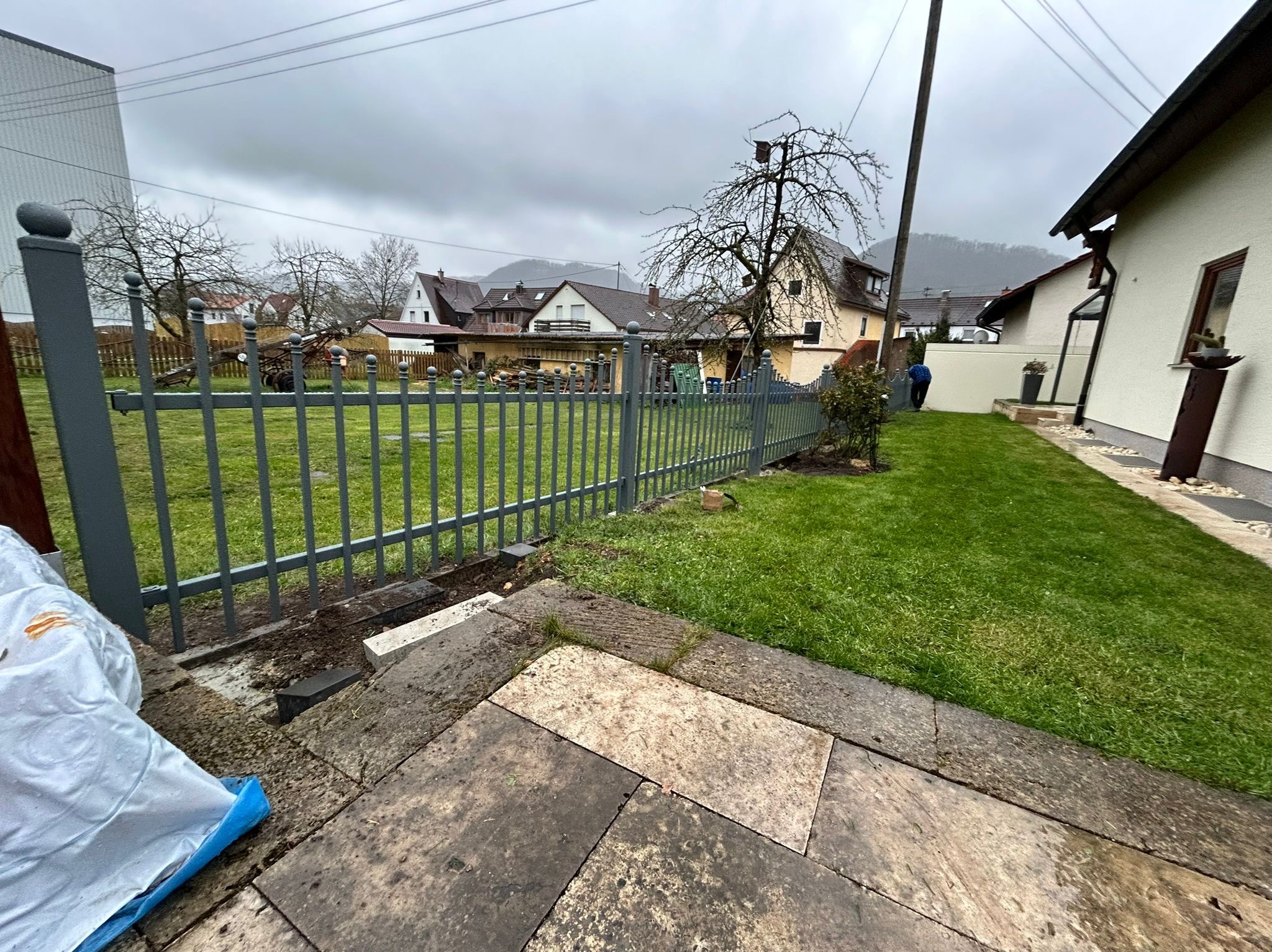 Szara, metalowa ogrodzenie posesji z kulistymi zakończeniami słupków, widoczne z perspektywy kamiennego chodnika w pochmurny dzień. W tle zabudowa mieszkalna.