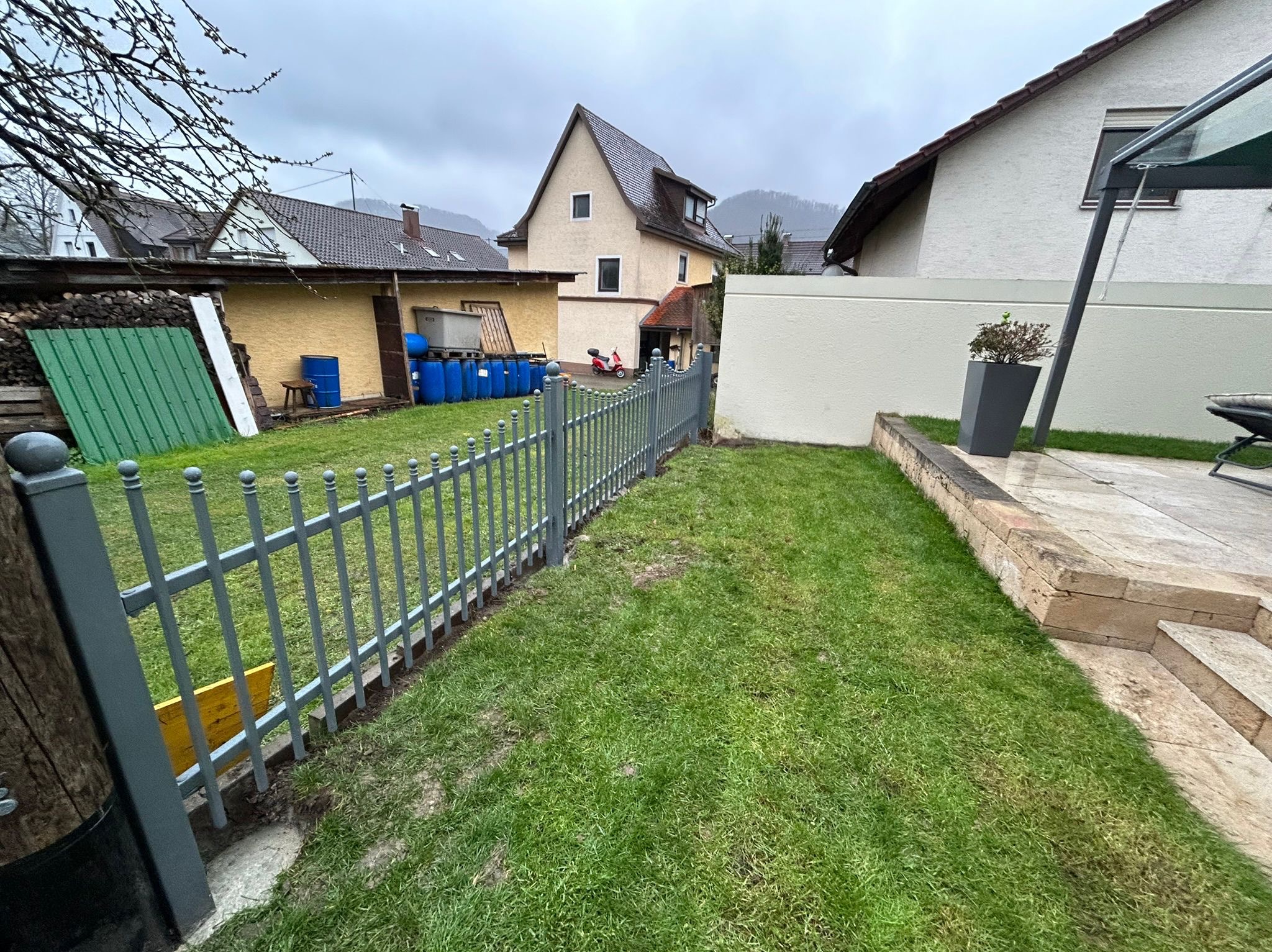 Szara metalowa palisada oddzielająca trawnik od zabudowań w pochmurny dzień. W tle budynki mieszkalne i wzgórza.