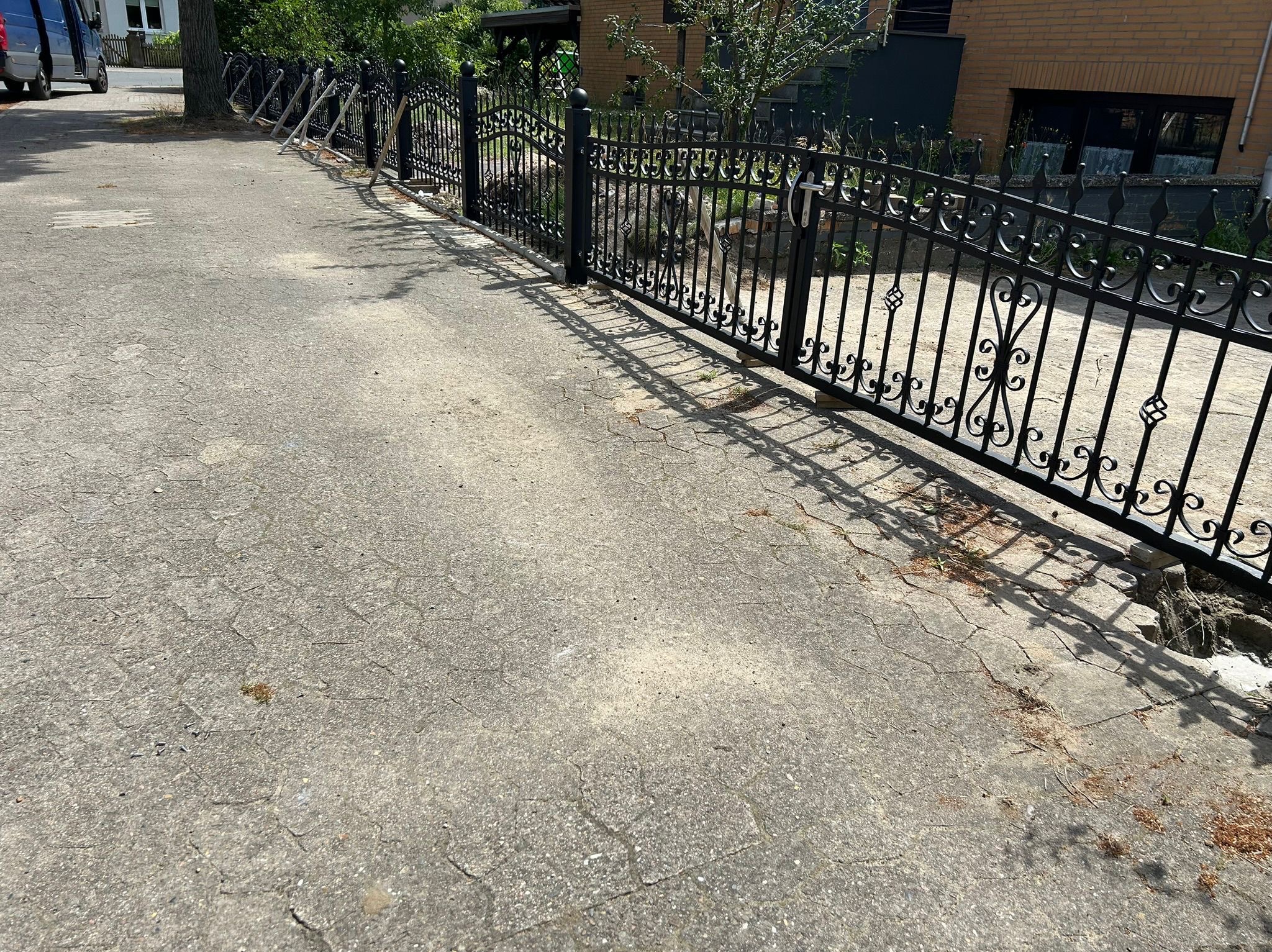 Czarny, dekoracyjny płot metalowy z bramą, zamontowany wzdłuż betonowej ścieżki. Widoczne podpory tymczasowe. Dom z cegły w tle, oświetlony słońcem.