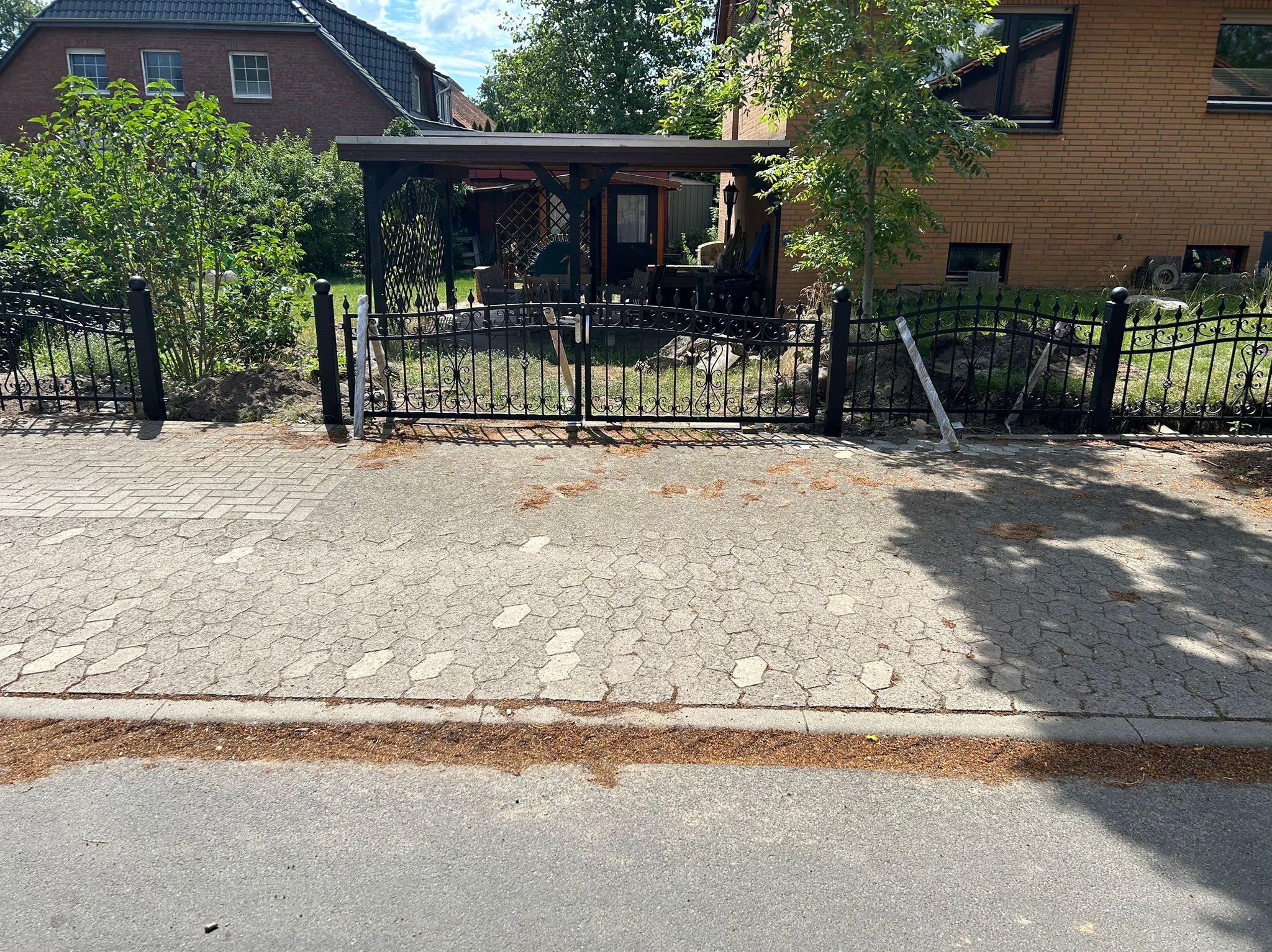 Czarna, metalowa brama z ozdobnymi elementami, częściowo zamontowana. Widoczne słupki w folii ochronnej, trawa i kostka brukowa. W tle dom z cegły i zadaszona konstrukcja.