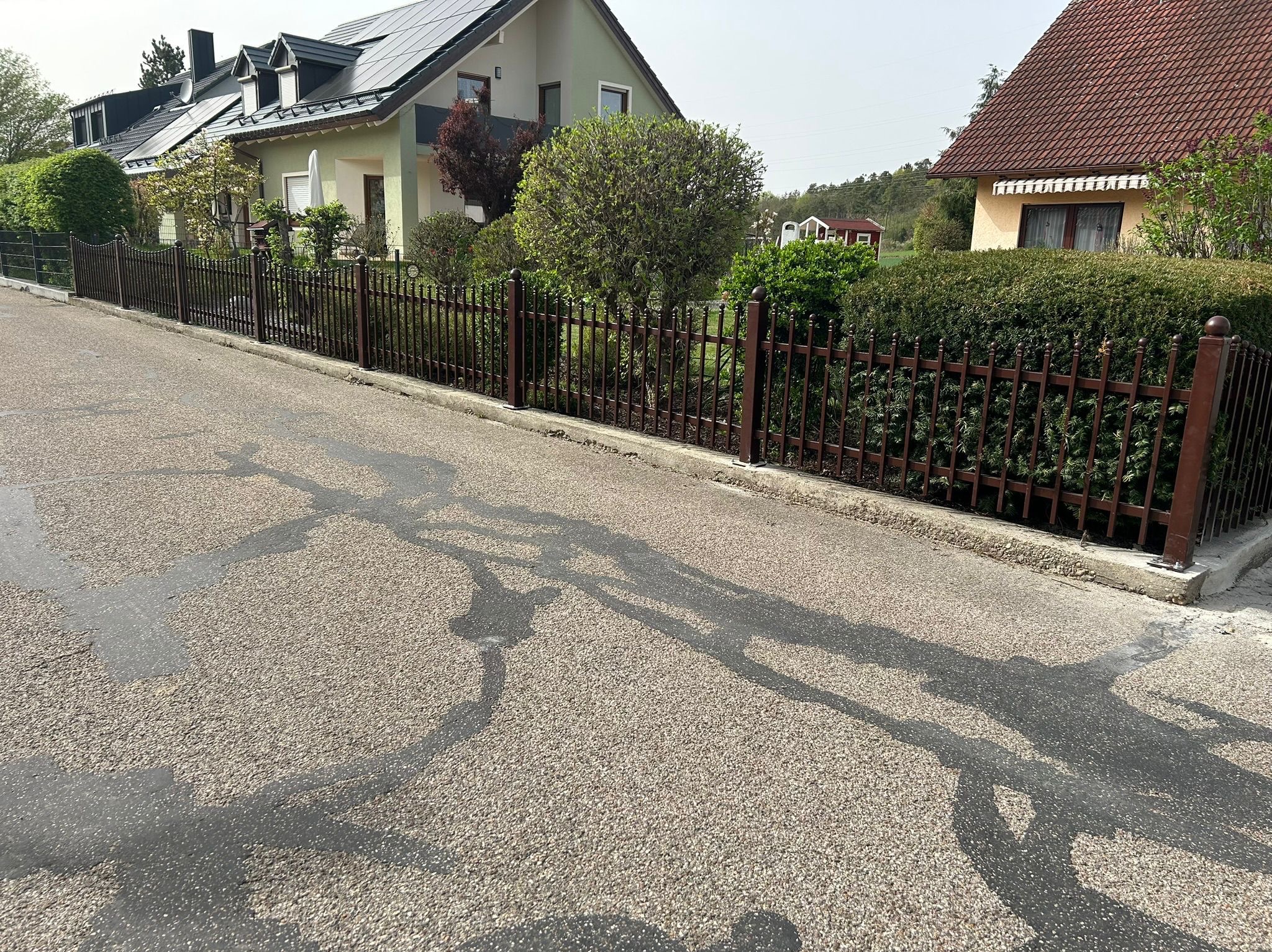 Brązowe ogrodzenie metalowe oddziela posesję od ulicy. Widoczne domy w tle i zieleń. Na asfalcie plamy wody. Słoneczny dzień.
