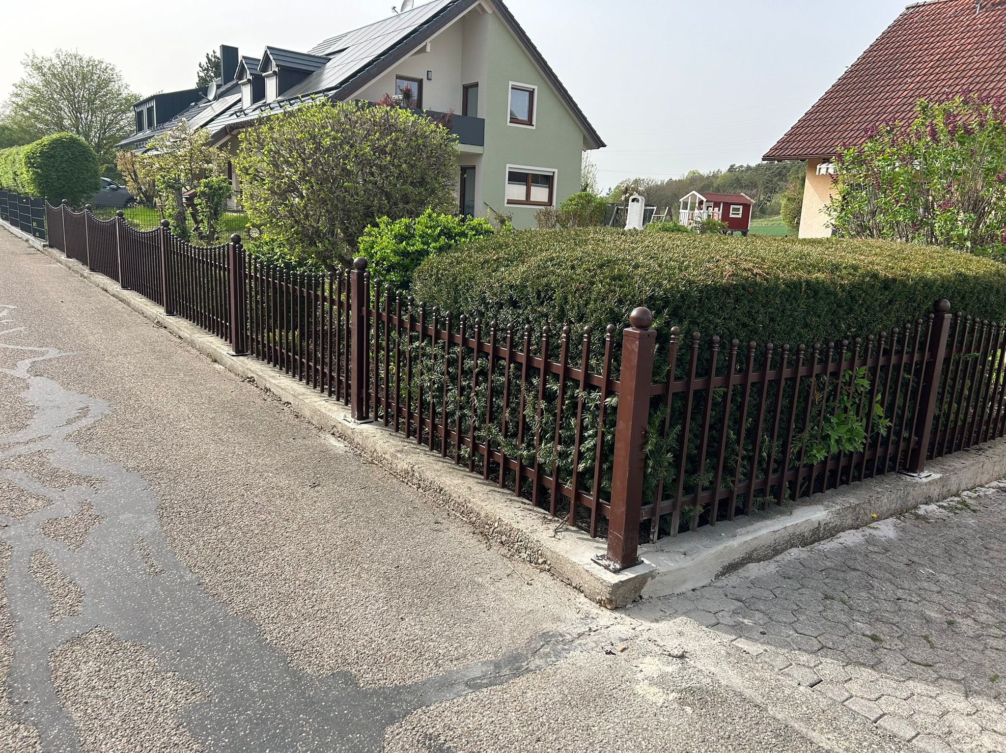 Ciemnobrązowe ogrodzenie metalowe z kulistymi zakończeniami słupków, oddzielające posesję od chodnika i ulicy. Za ogrodzeniem gęsta roślinność i dom.