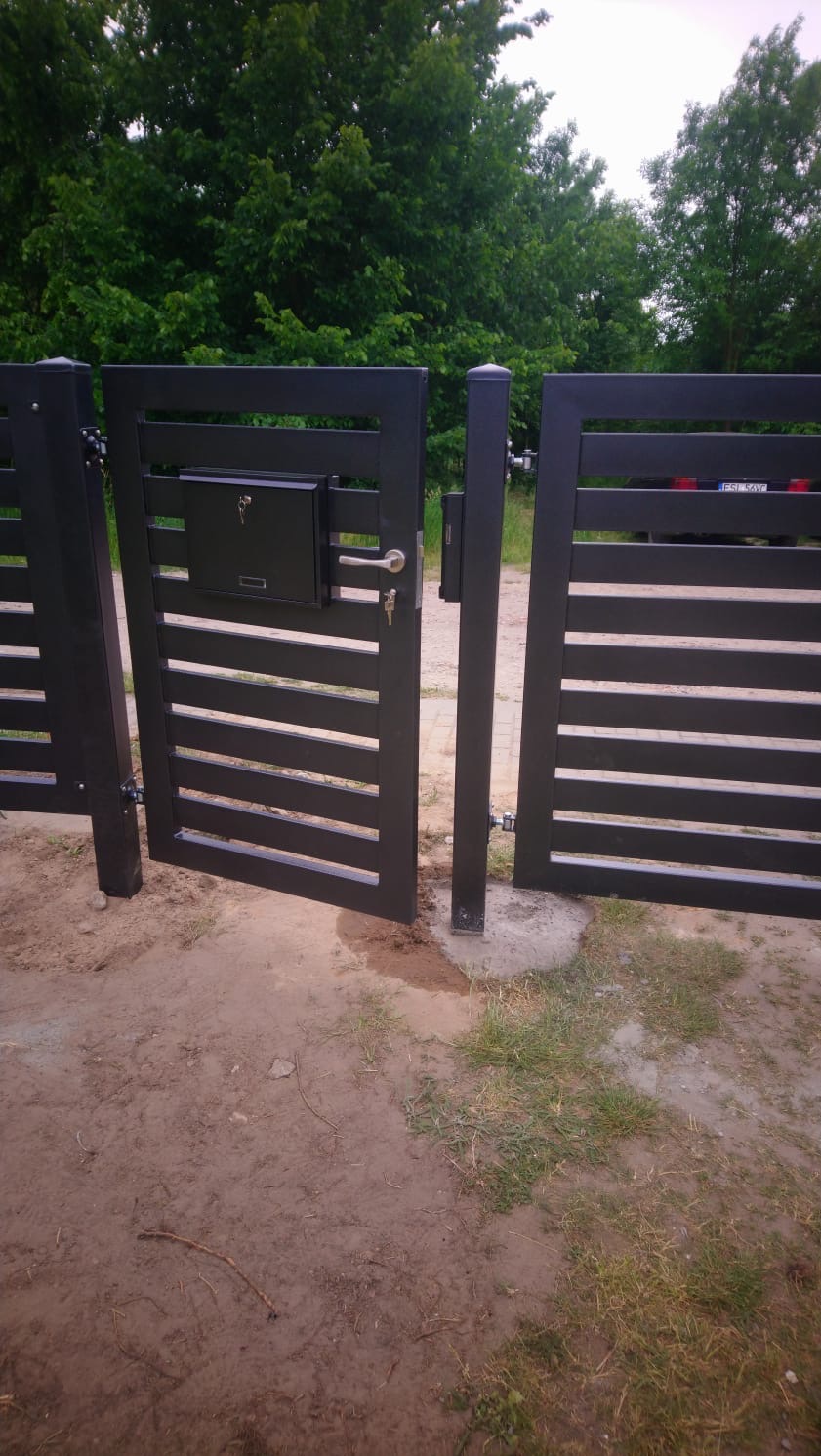 Nowoczesna, grafitowa brama wjazdowa z poziomymi szczeblami i skrzynką na listy, otwierana na posesję z zielenią w tle. Widoczne zawiasy i zamek.