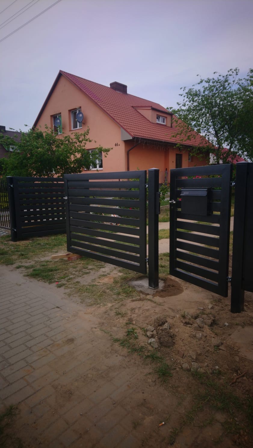 Nowoczesna, grafitowa brama posesyjna z poziomymi panelami i skrzynką na listy, na tle domu z czerwoną dachówką i zieleni. Widok z zewnątrz.