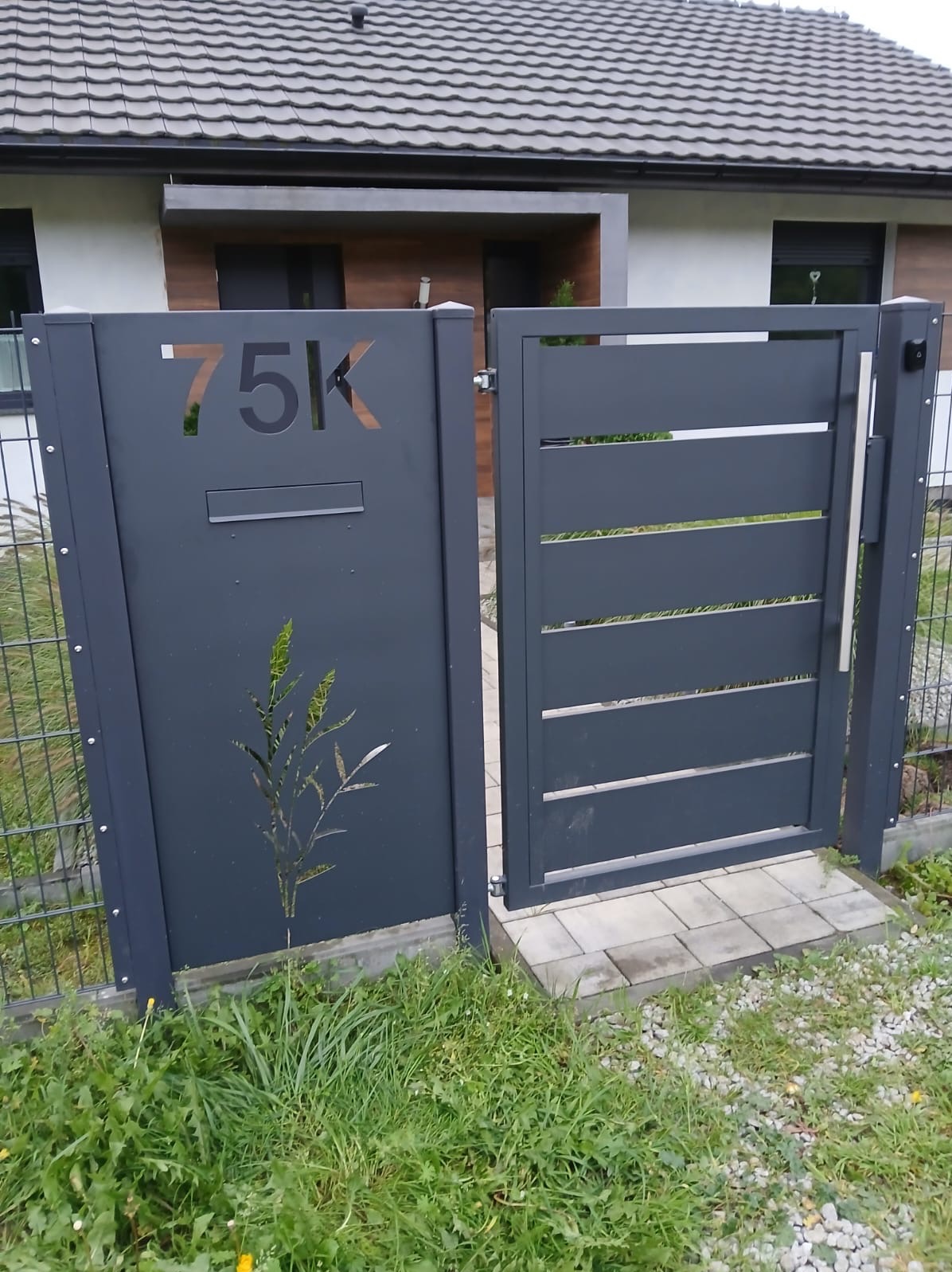 Nowoczesna, antracytowa furtka z numerem domu '75K' wyciętym laserowo i motywem roślinnym. Furtka z poziomymi szczeblami, na tle domu z szarą dachówką.