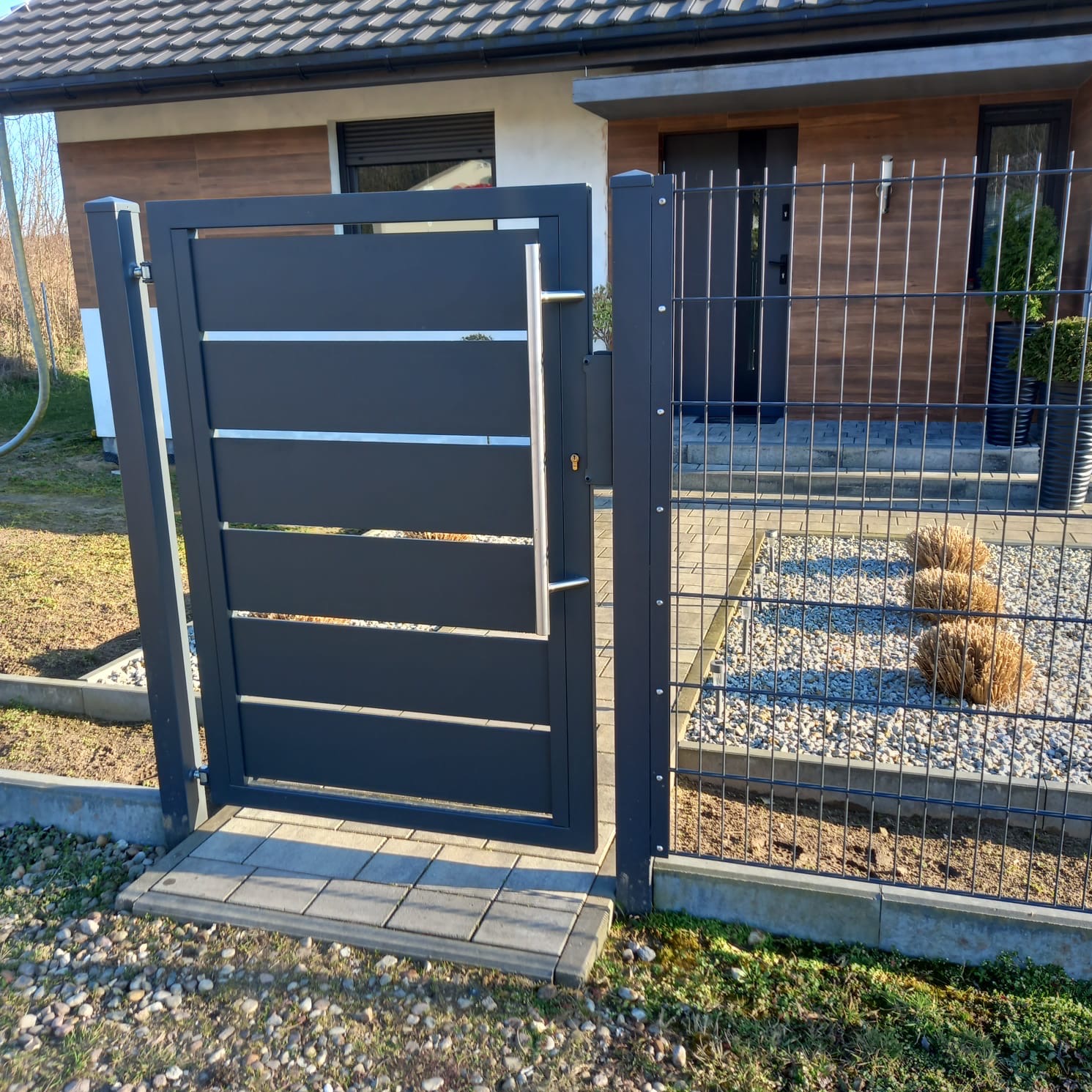 Nowoczesna, antracytowa furtka z poziomymi panelami i srebrnymi akcentami, wkomponowana w ogrodzenie z siatki na tle domu z drewnianą elewacją.