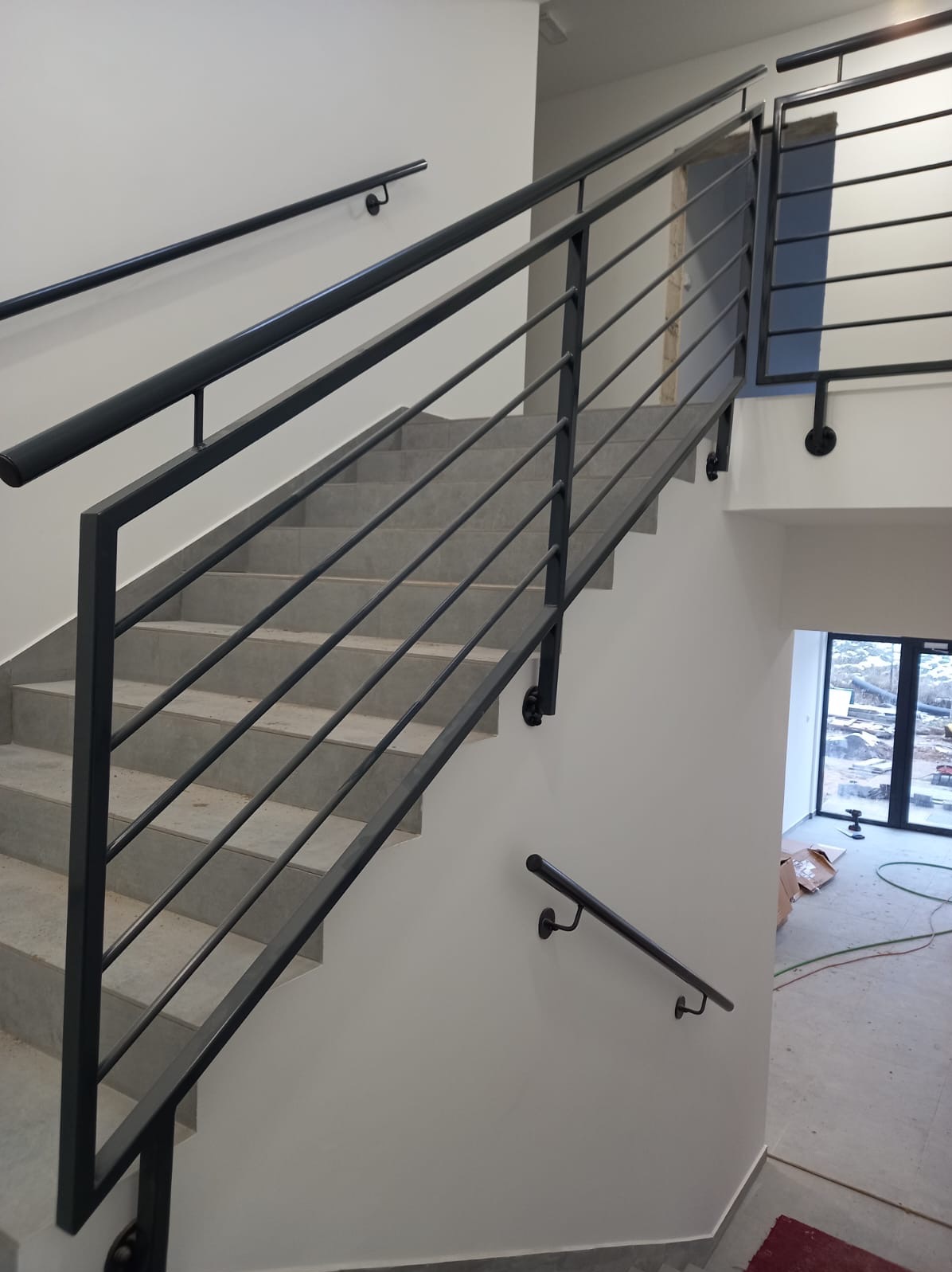 Nowoczesne schody z metalową balustradą w minimalistycznym stylu. Szare stopnie, czarne, poziome pręty balustrady. Widok z dołu schodów, w tle okno z widokiem na budowę.