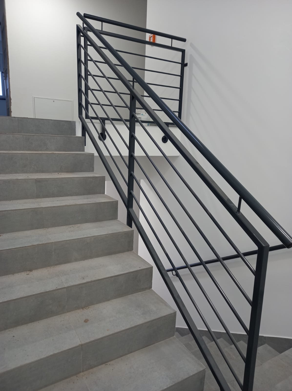 Nowoczesne schody z szarymi płytkami i minimalistyczną, metalową balustradą w kolorze grafitowym. Widok z dołu, podkreślający geometrię konstrukcji i surowy charakter wnętrza.