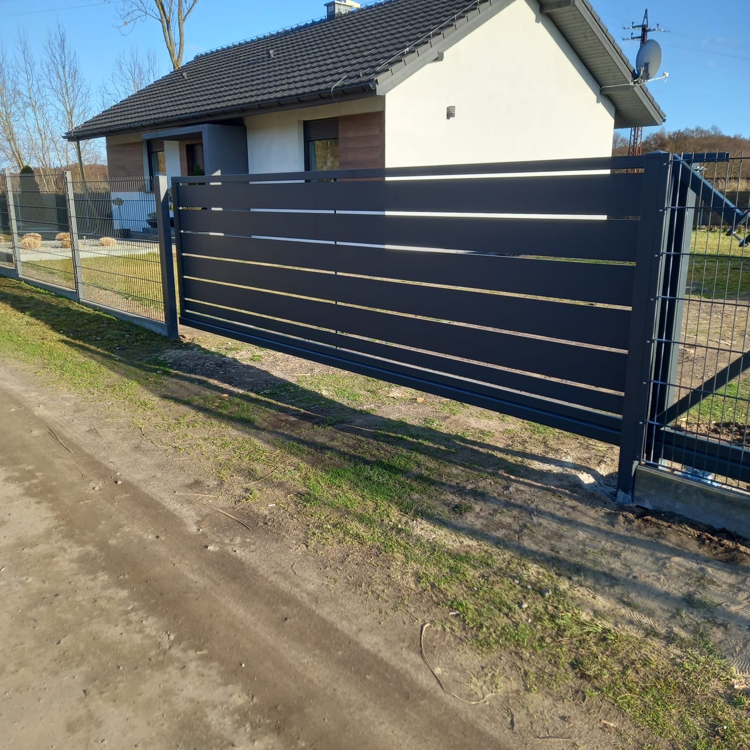 Nowoczesna, przesuwna brama w kolorze antracytowym z poziomymi panelami, dopasowana do ogrodzenia z siatki na tle domu z ciemnym dachem.