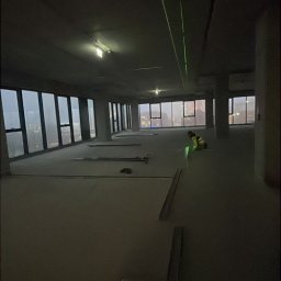 PKB GROUP SPÓŁKA Z OGRANICZONĄ ODPOWIEDZIALNOŚCIĄ - Wnętrze w trakcie prac wykończeniowych. Widoczne profile, poziomica laserowa wyznaczająca płaszczyzny. W tle okna z widokiem na miasto i pracownik w kamizelce.