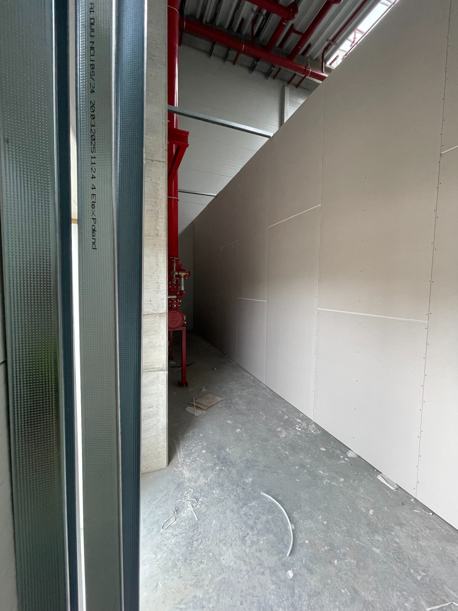 Montaż ścian działowych z płyt gipsowo-kartonowych w surowym wnętrzu. Widoczne metalowe profile i czerwona instalacja przeciwpożarowa.