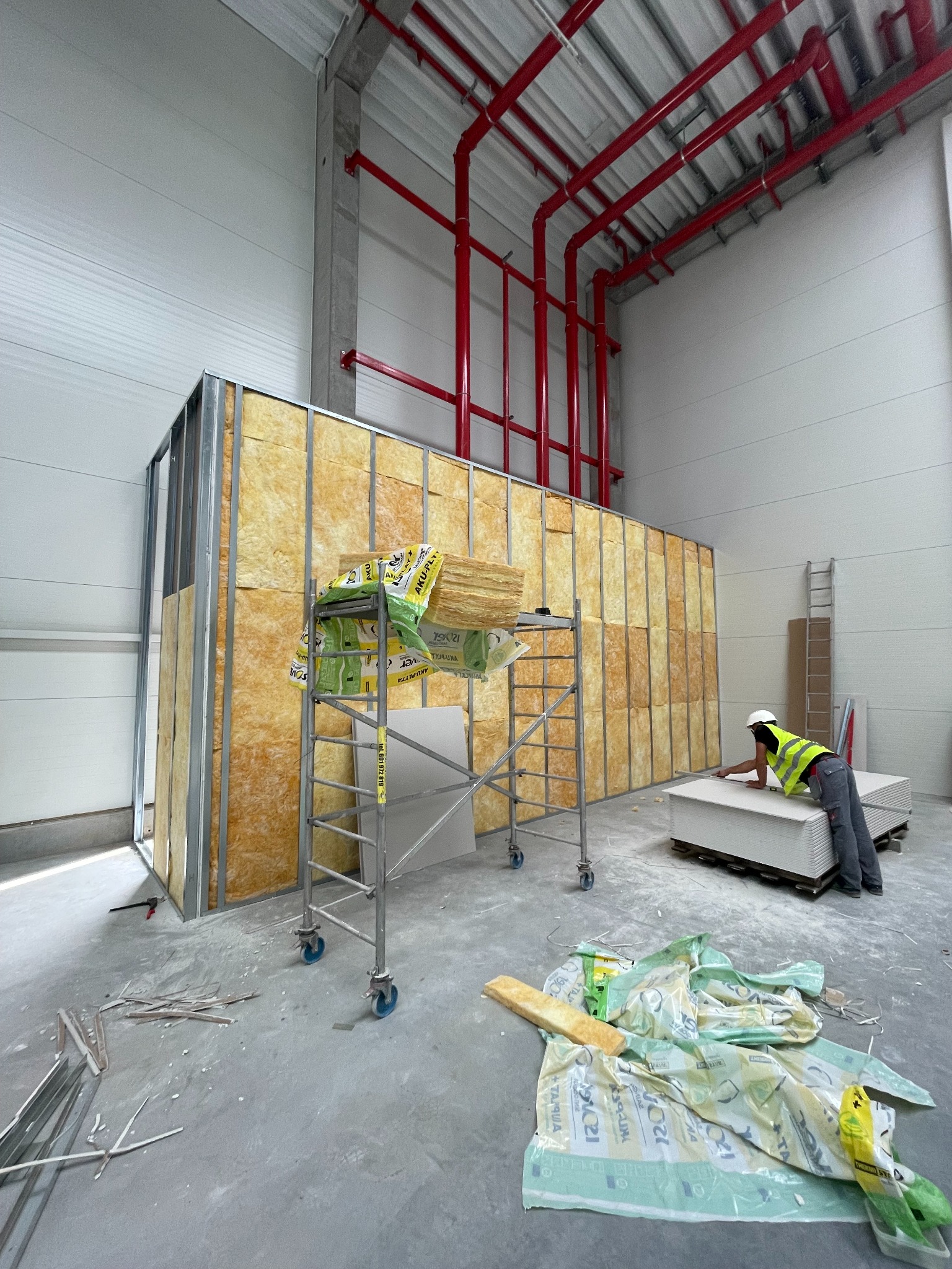 Montaż ścianki działowej z izolacją z wełny mineralnej w hali. Widoczne profile, płyty gipsowe i rusztowanie. Pracownik w kasku i kamizelce mierzy płytę.